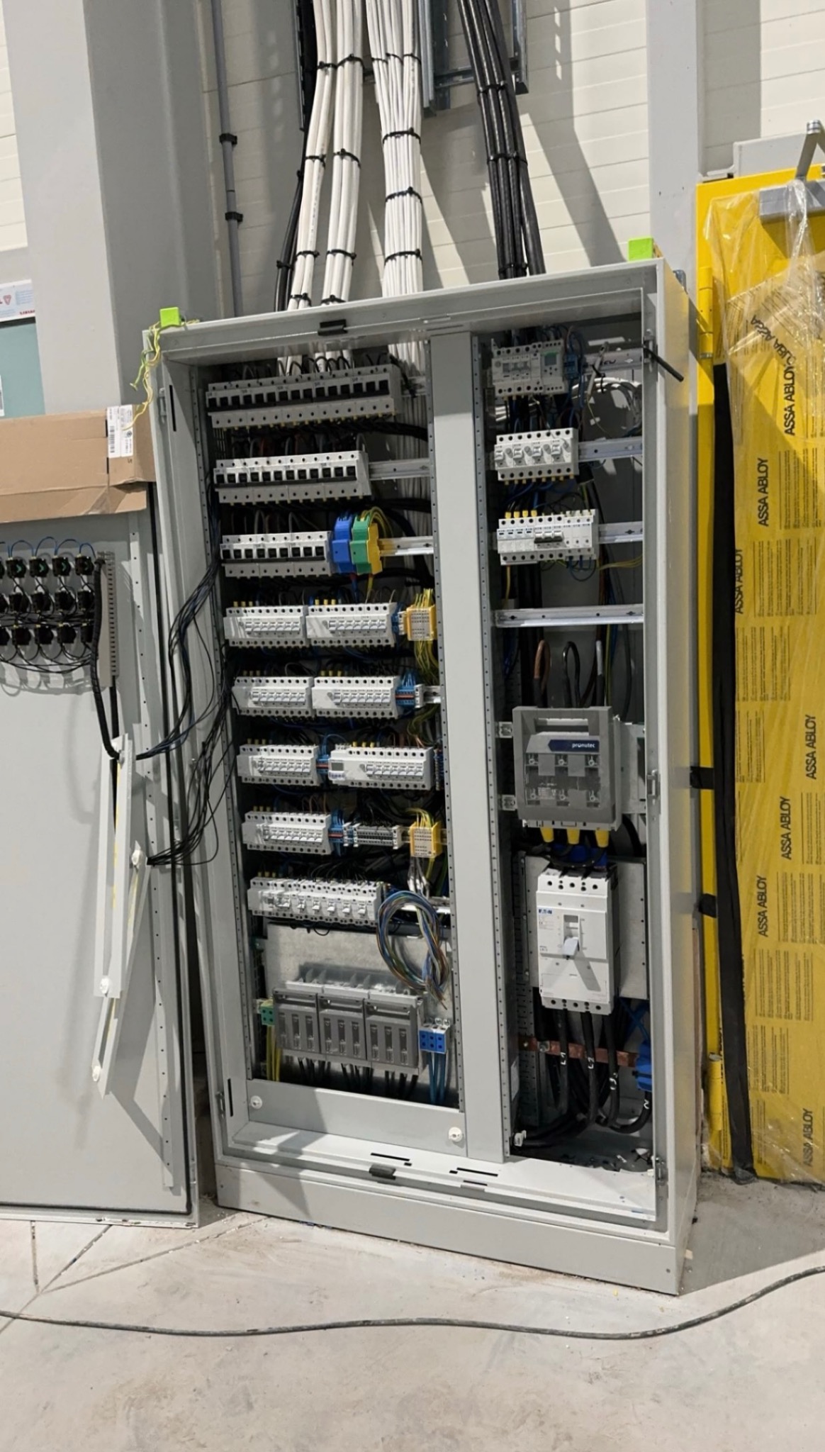 Otwarta szafa sterownicza z rozdzielnicą elektryczną, widoczne bezpieczniki, kable i przewody, obok zapakowane płyty izolacyjne.