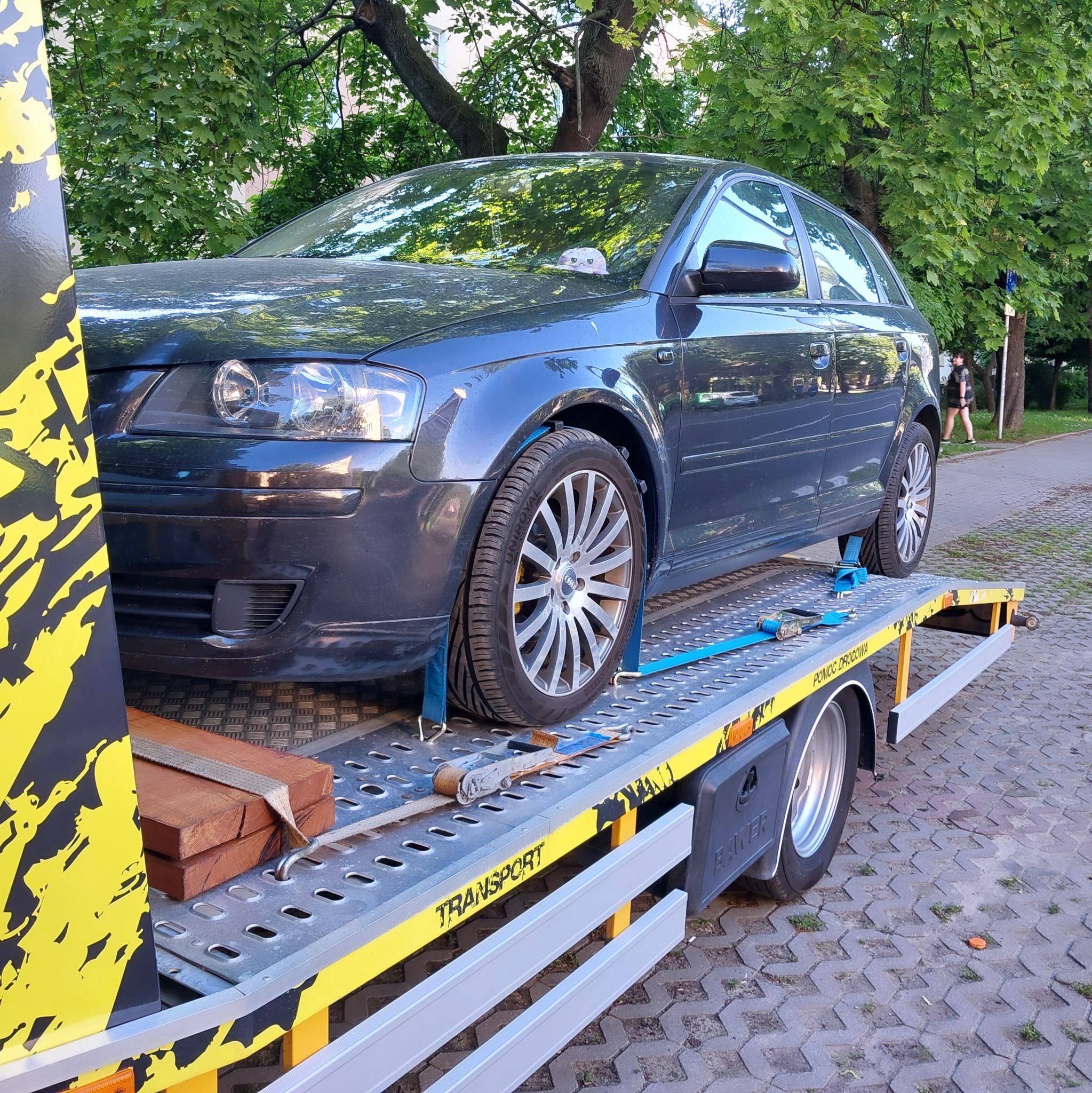 Ciemnoszary samochód osobowy marki Audi, zabezpieczony niebieskimi pasami transportowymi, przewożony na żółto-szarej lawecie na tle zielonych drzew.
