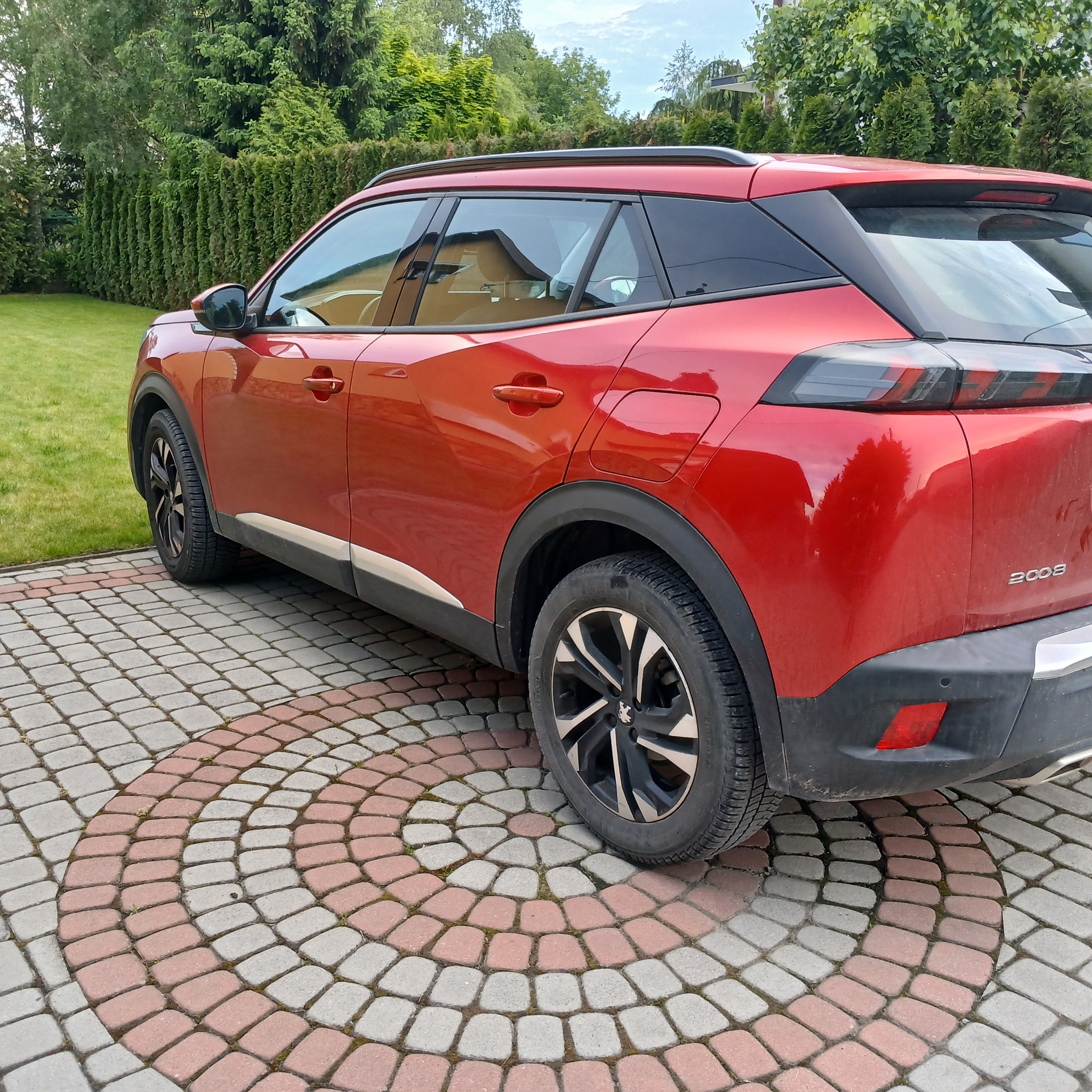 Czerwony Peugeot 2008 na ozdobnej kostce brukowej, zieleń w tle. Widok z tyłu, lekko z boku, podkreśla nowoczesny design i kolor auta.