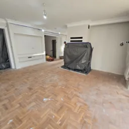 Pok&oacute;j z parkietem w jodełkę przed renowacją, zabezpieczone meble folią, widoczne ślady po szpachlowaniu na podłodze.