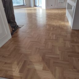 Cyklinpro.pl - Ułożony parkiet w jodełkę francuską, widoczne zabezpieczone folią okna i grzejnik na ścianie, fragment osoby w kombinezonie ochronnym.