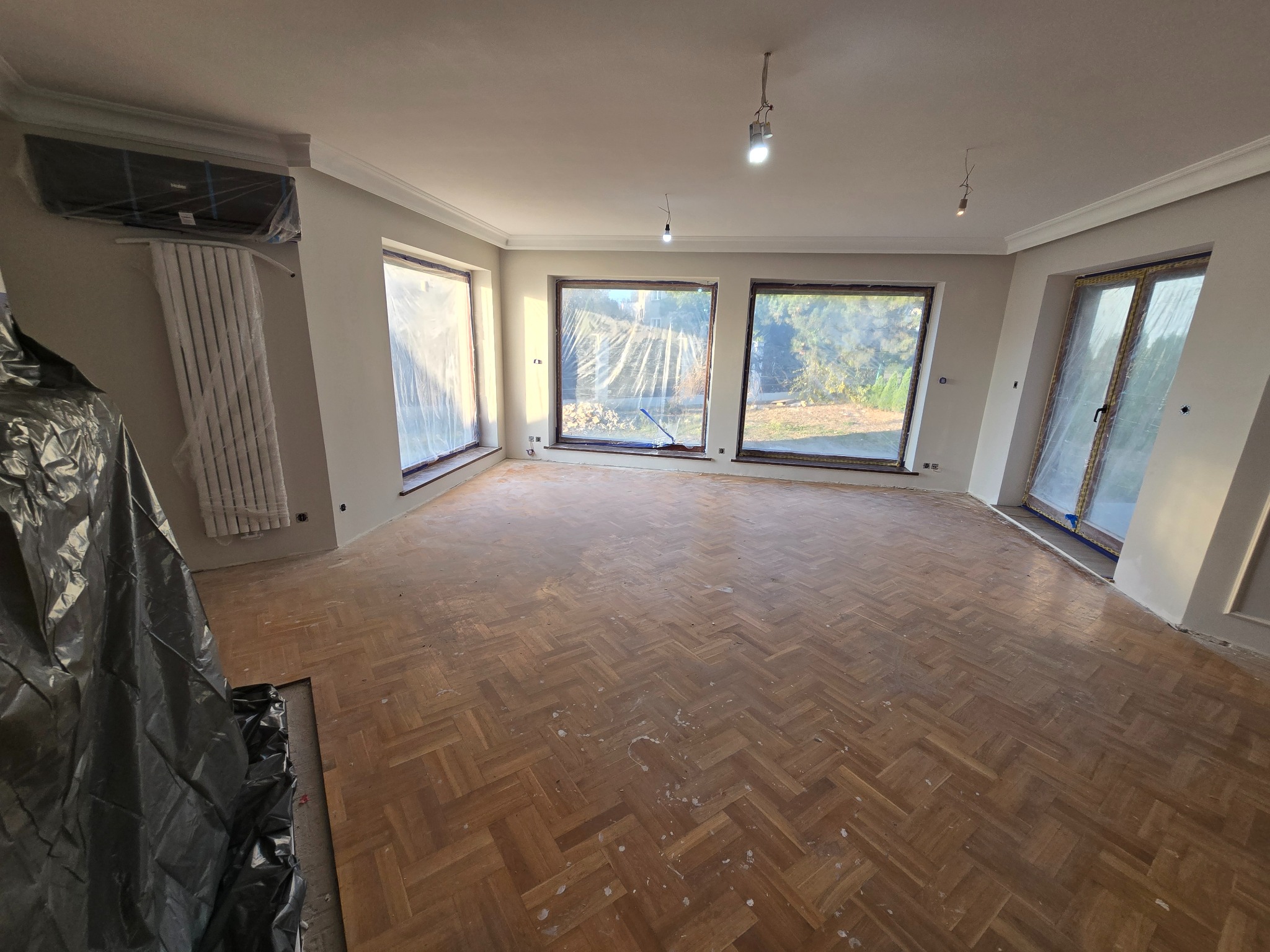 Pokój z parkietem ułożonym w jodełkę, przygotowany do renowacji, okna zabezpieczone folią, widoczny grzejnik i klimatyzator.