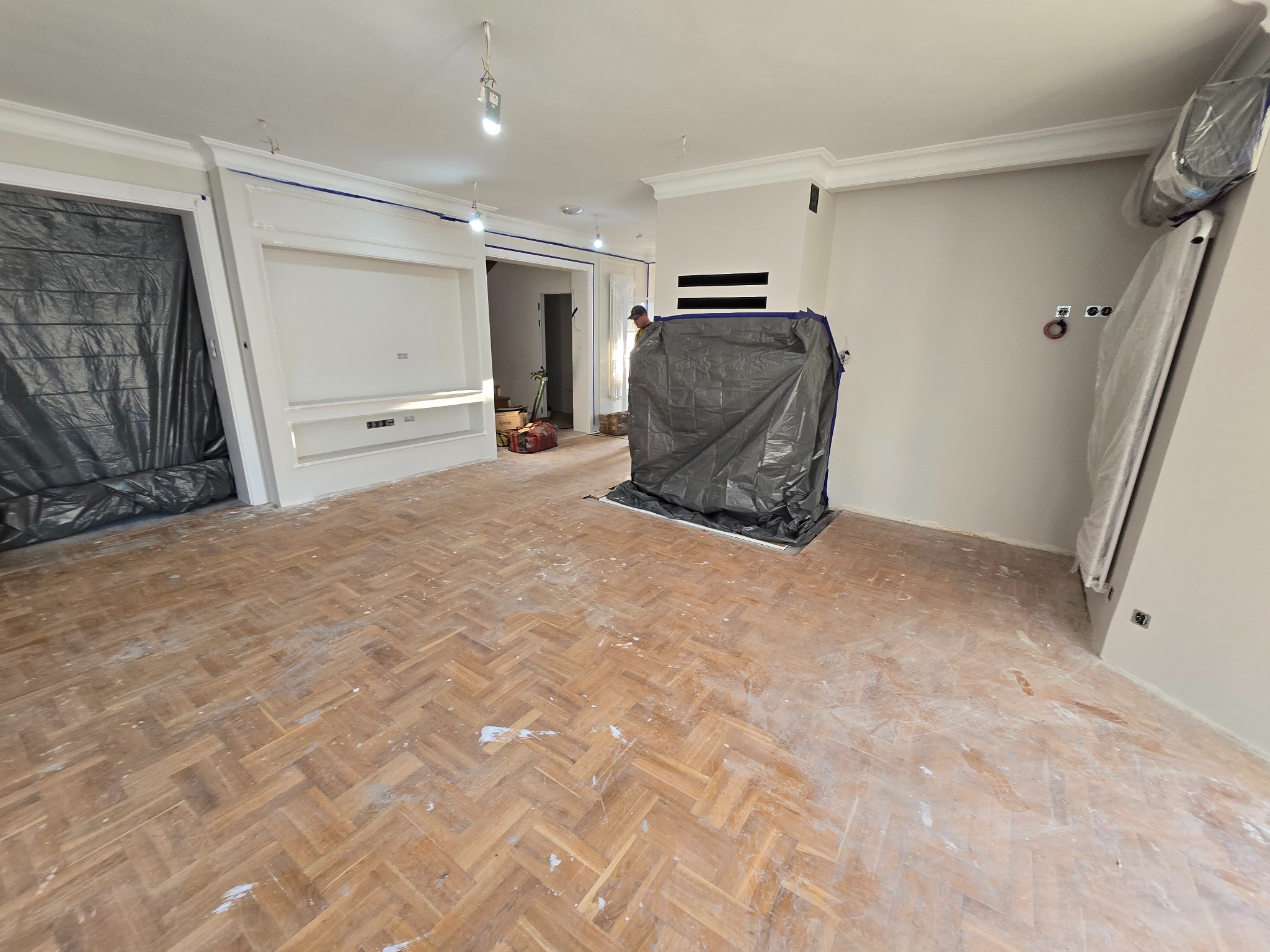 Pokój z parkietem w jodełkę przed renowacją, zabezpieczone meble folią, widoczne ślady po szpachlowaniu na podłodze.