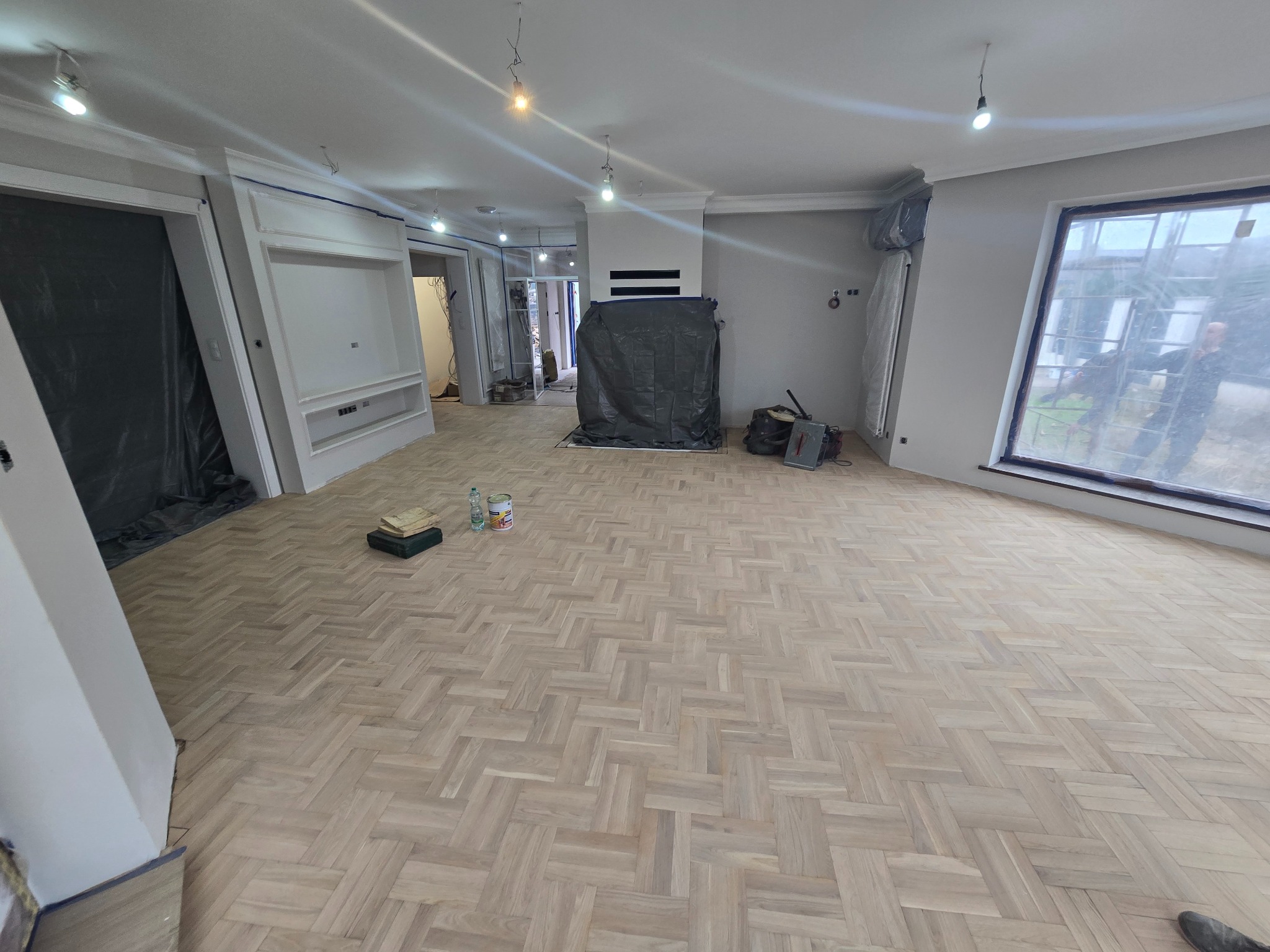 Nowo ułożony parkiet w jodełkę w przestronnym salonie w Świdnicy, z widocznymi przygotowaniami do dalszych prac wykończeniowych, takimi jak zabezpieczone meble i ściany.