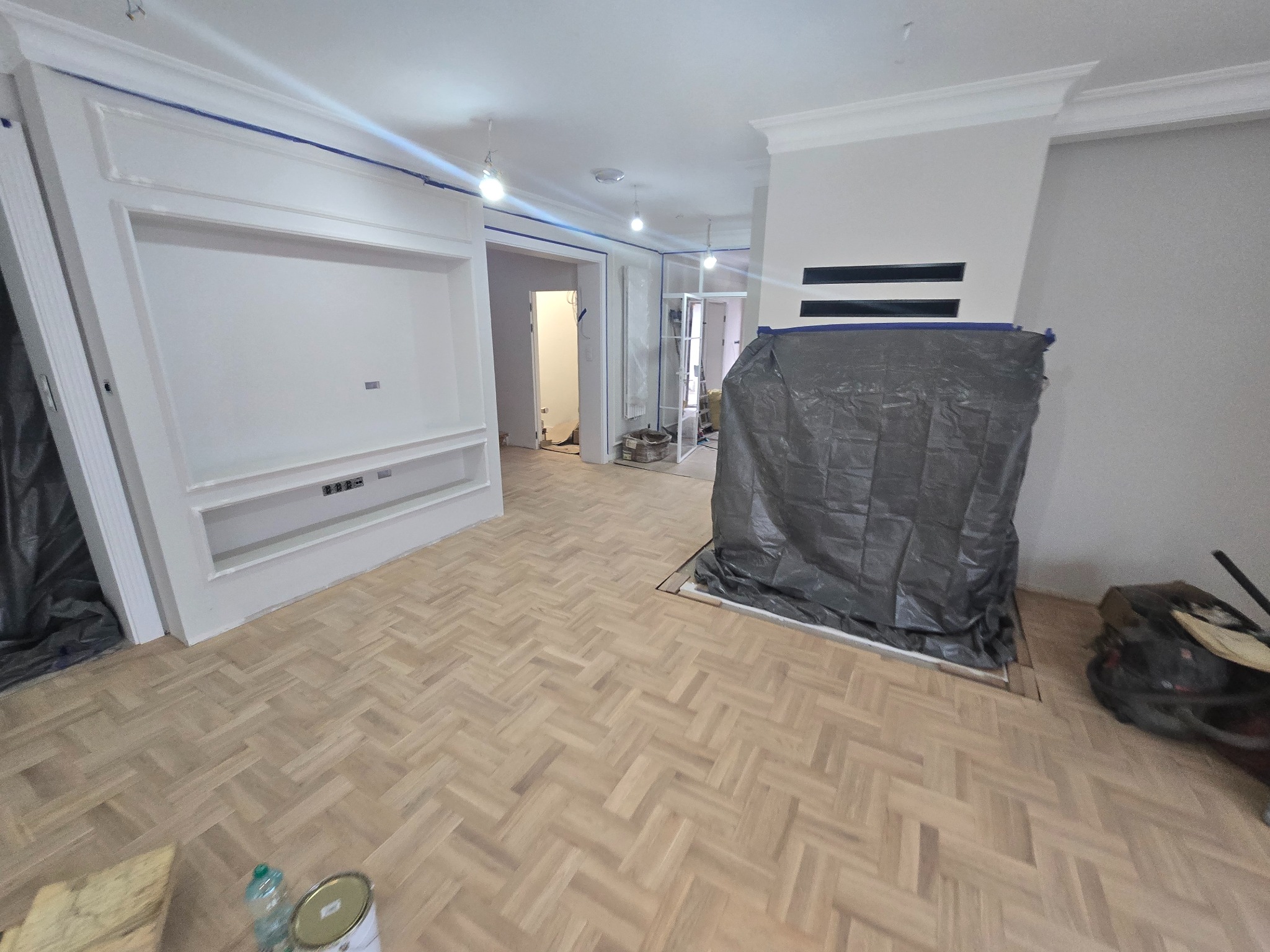 Ułożony parkiet w jodełkę francuską w jasnym odcieniu, z zabezpieczonym folią kominkiem i ścianami przygotowanymi do malowania, widoczna puszka farby i butelka wody mineralnej.