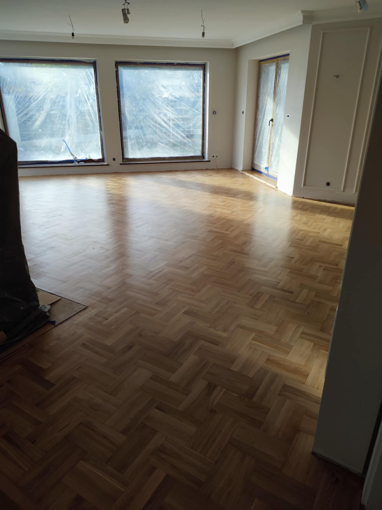 Parkiet ułożony w jodełkę w przestronnym, jasnym pomieszczeniu z oknami zabezpieczonymi folią ochronną podczas prac remontowych.