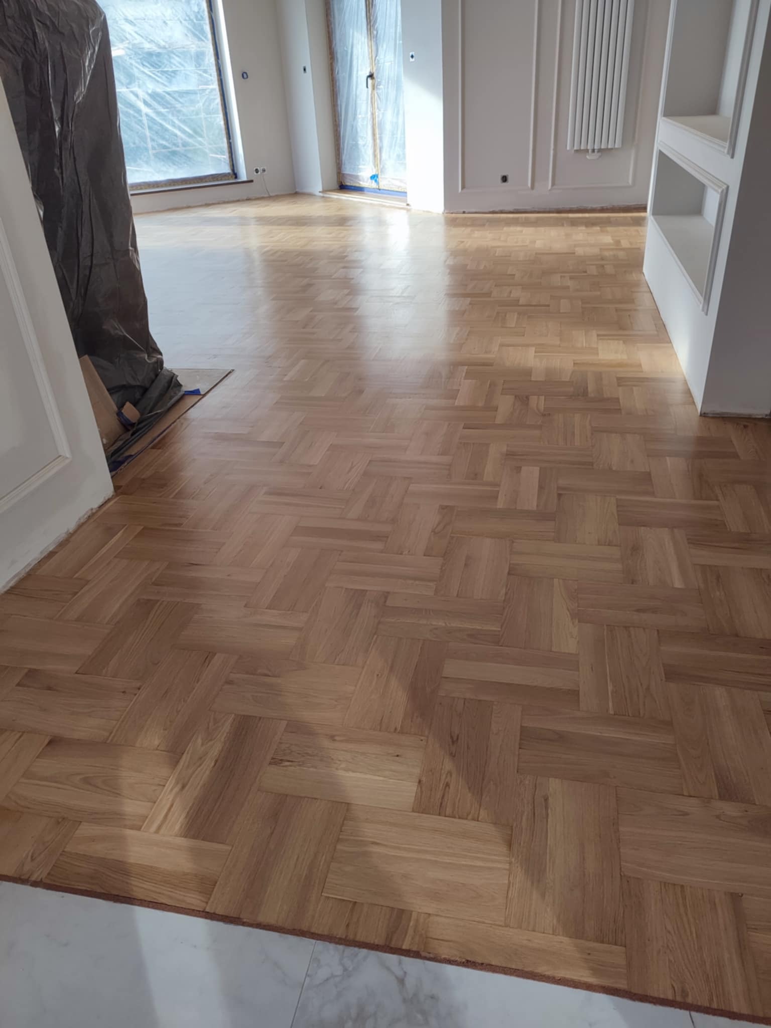 Ułożony parkiet w jodełkę francuską, widoczne zabezpieczone folią okna i grzejnik na ścianie, fragment osoby w kombinezonie ochronnym.