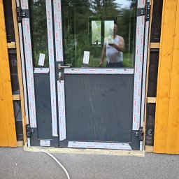 KW.BRUK - Montaż dwuskrzydłowych drzwi zewnętrznych z czarną ramą, widoczne tymczasowe zabezpieczenia i odblask sylwetki w szybie.