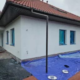 Biała elewacja domu jednorodzinnego w trakcie ocieplania, okna zabezpieczone folią, widoczne plandeki ochronne na ziemi i fragment dachu z czerwonej dachówki.