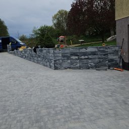 KW.BRUK - Szara kostka brukowa ułożona na podjeździe, z niskim murem oporowym z ciemniejszych kostek, w tle zaparkowany niebieski bus i betoniarka.