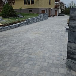 KW.BRUK - Ułożona szara kostka brukowa prowadząca do domu z beżową elewacją i czerwoną dachówką, z widocznym fragmentem murku z kostki i stosem nieułożonych elementów po prawej stronie.