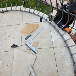 KW.BRUK - Montaż płytek na zakrzywionym balkonie z metalową balustradą; widoczne narzędzia: nóż, kątownik, nożyczki i miarka składana; pracownik w rękawicach ochronnych przycina płytkę.