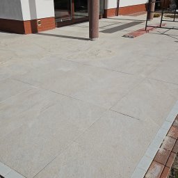 KW.BRUK - Jasnoszara płyta tarasowa z obrzeżem z czerwonej cegły i szarej kostki, widok z góry na tle domu z brązowymi oknami i kolumną.