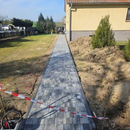 KW.BRUK - Świeżo ułożony chodnik z szarej kostki brukowej prowadzący wzdłuż domu z pomarańczowym dachem, zabezpieczony taśmą ostrzegawczą, z widoczną stertą ziemi i dwoma osobami w oddali.