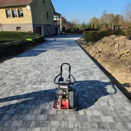 Świeżo ułożona, szara kostka brukowa prowadząca do domu, z zagęszczarką w centrum kadru i widoczną ziemią po bokach ścieżki.