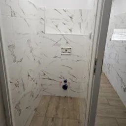 Nowoczesna, wąska łazienka z białymi płytkami imitującymi marmur i drewnopodobną podłogą, widoczne elementy instalacji hydraulicznej przed montażem armatury.