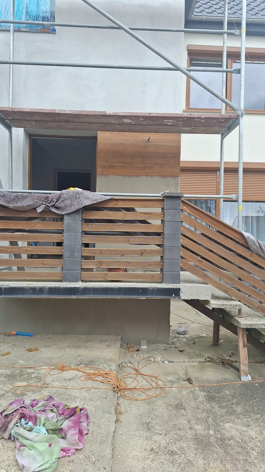 Elewacja domu w trakcie prac wykończeniowych: ocieplenie, drewniana okładzina, balustrada z drewnianymi elementami i betonowymi słupkami, rusztowanie. Widoczne narzędzia i materiały budowlane.