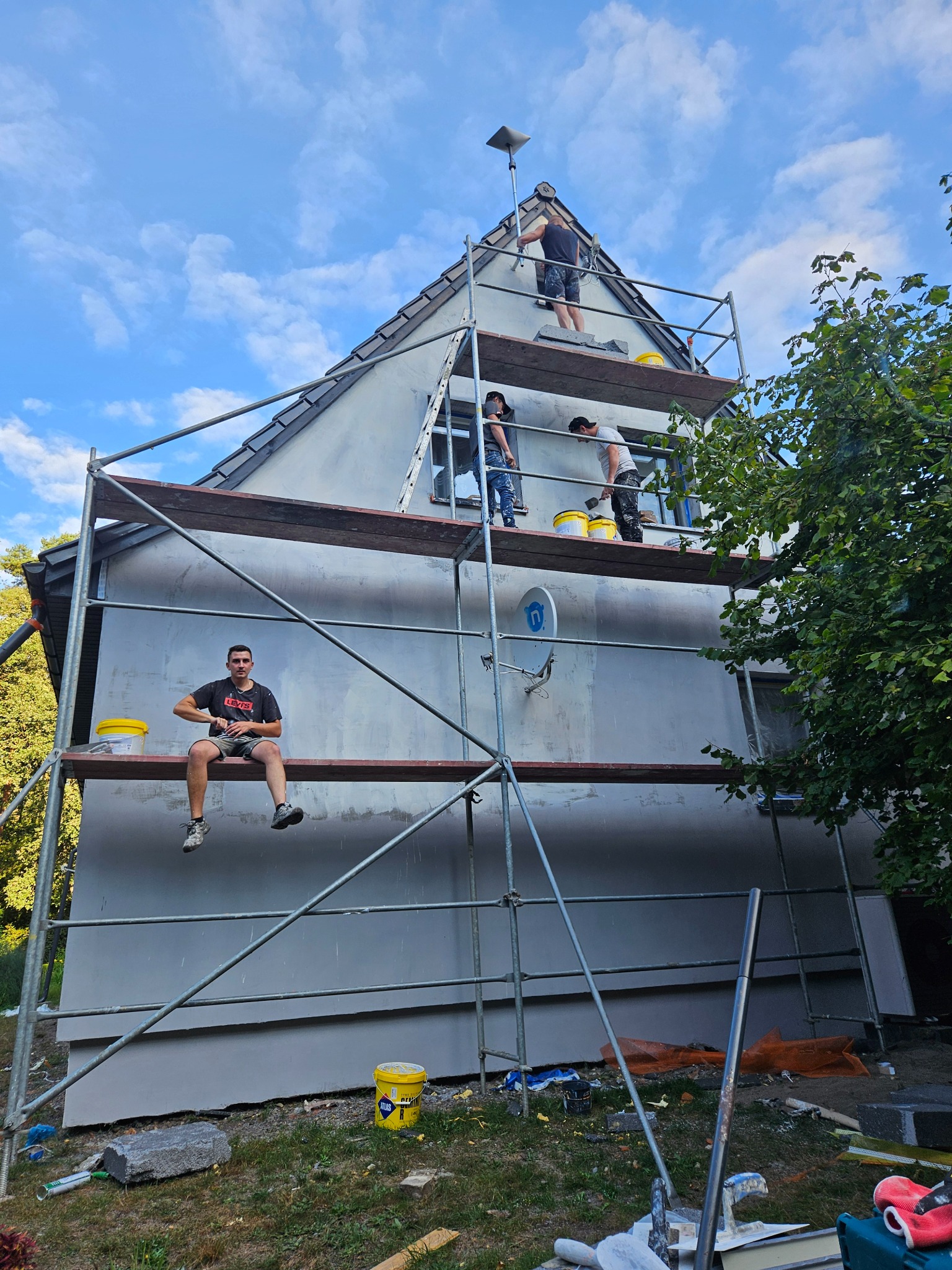Ocieplanie elewacji domu jednorodzinnego: ekipa na rusztowaniu, jeden pracownik siedzi na rusztowaniu z wiadrem, pozostali pracują nad nałożeniem tynku. Widoczne narzędzia i materiały budowlane.