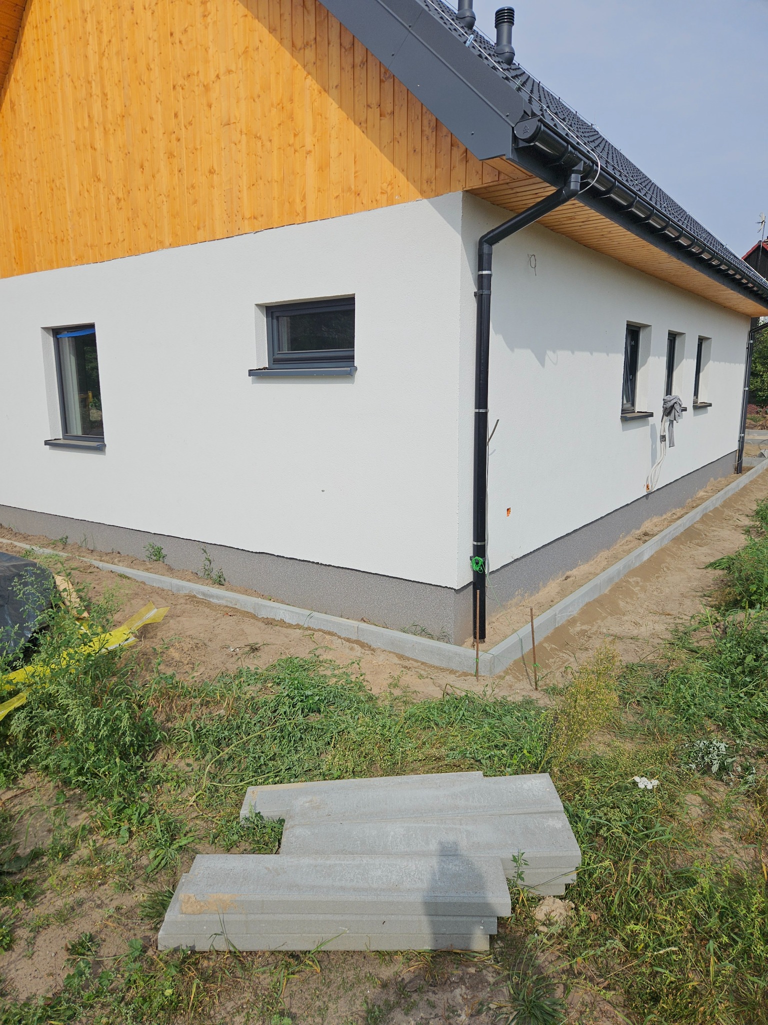 Nowoczesny dom jednorodzinny z białymi ścianami, drewnianym szczytem i szarą podmurówką, widoczne ułożone obrzeża trawnikowe przed budynkiem, teren wokół w trakcie prac wykończeniowych.