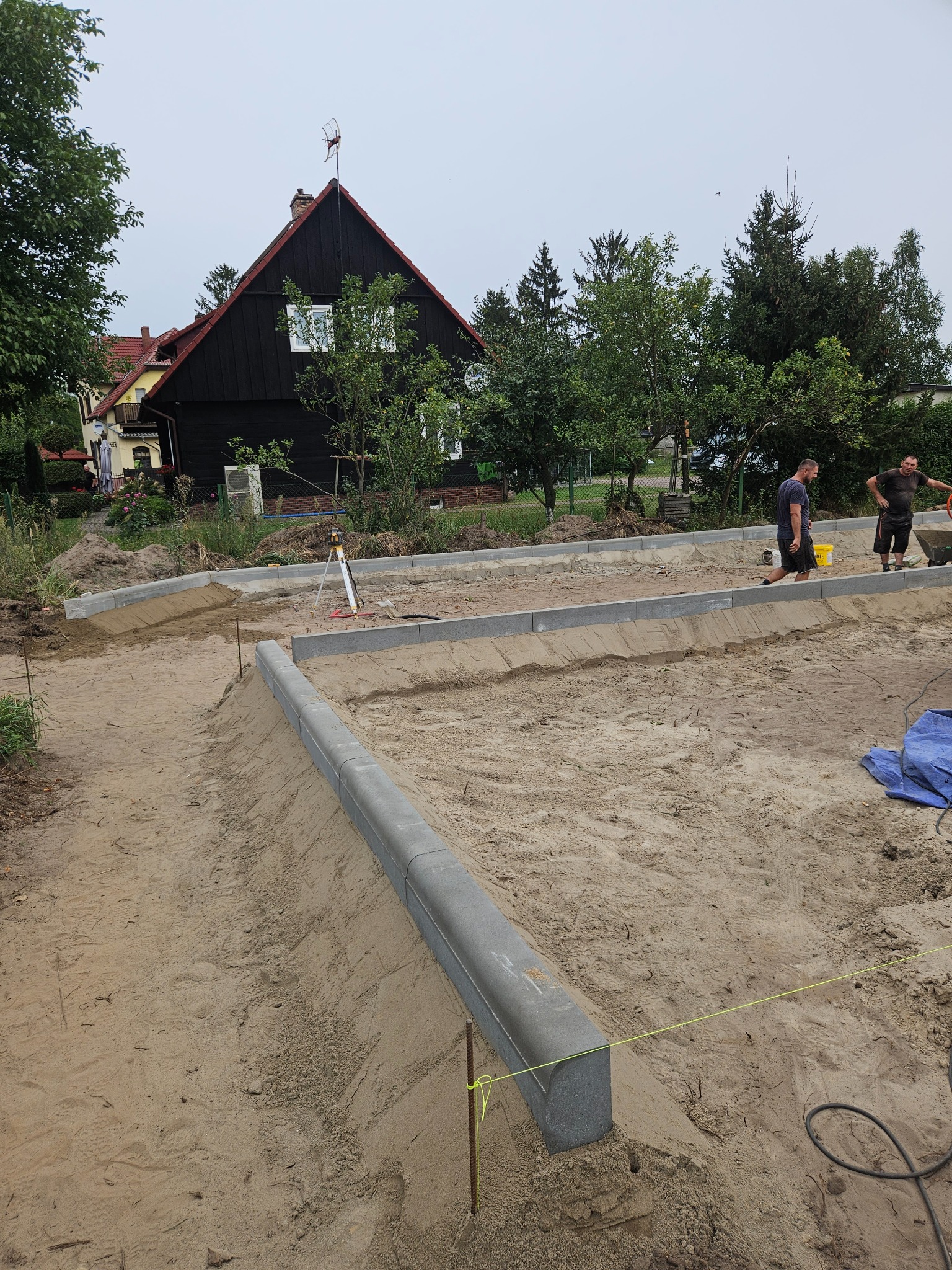 Przygotowanie terenu pod przyszły chodnik z obrzeżami, widoczny niwelator optyczny i dwaj pracownicy w tle.