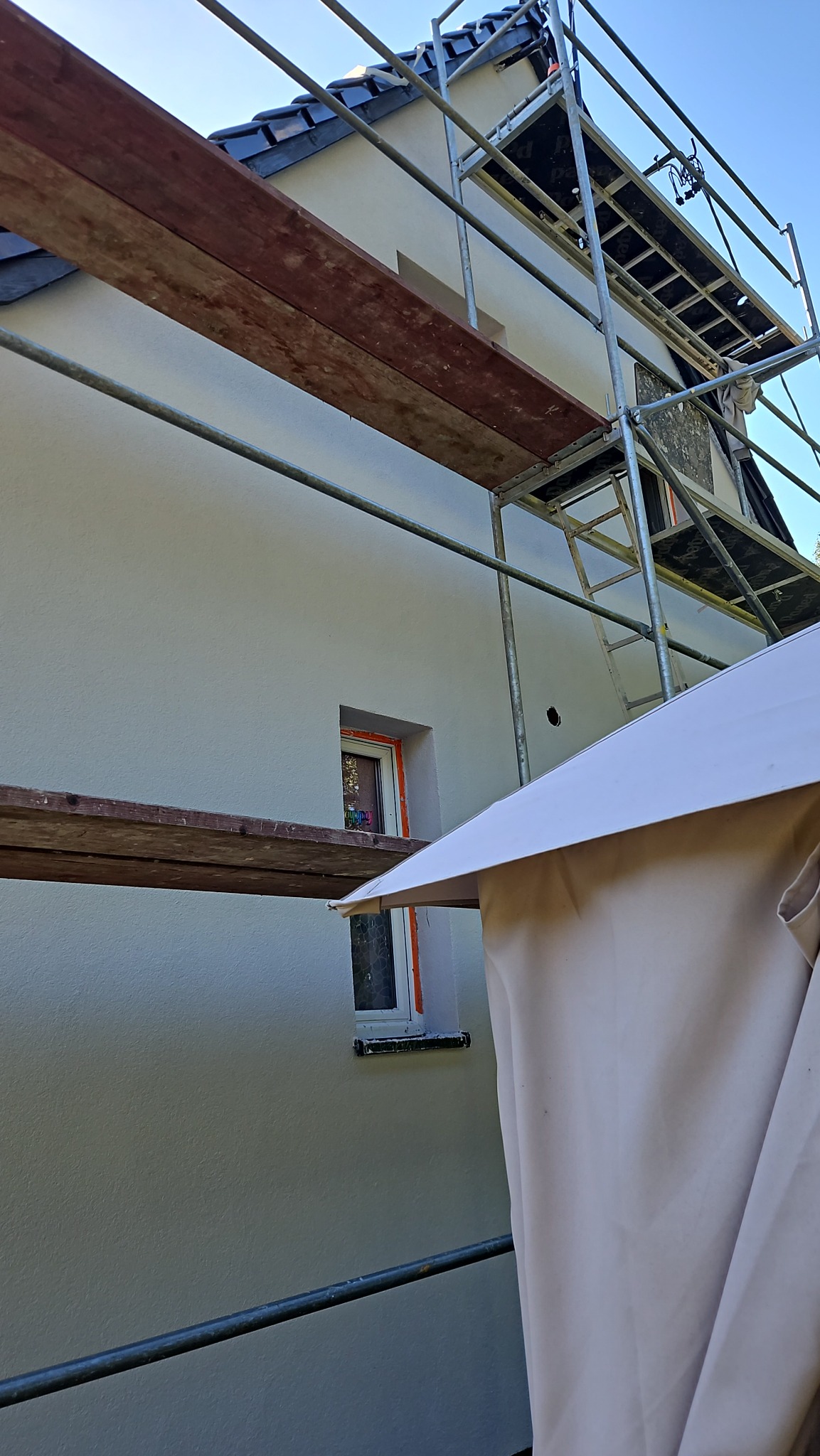 Elewacja budynku w trakcie ocieplania, widoczne rusztowanie, zabezpieczone okno z pomarańczową ramą, świeżo nałożony tynk w jasnym odcieniu.