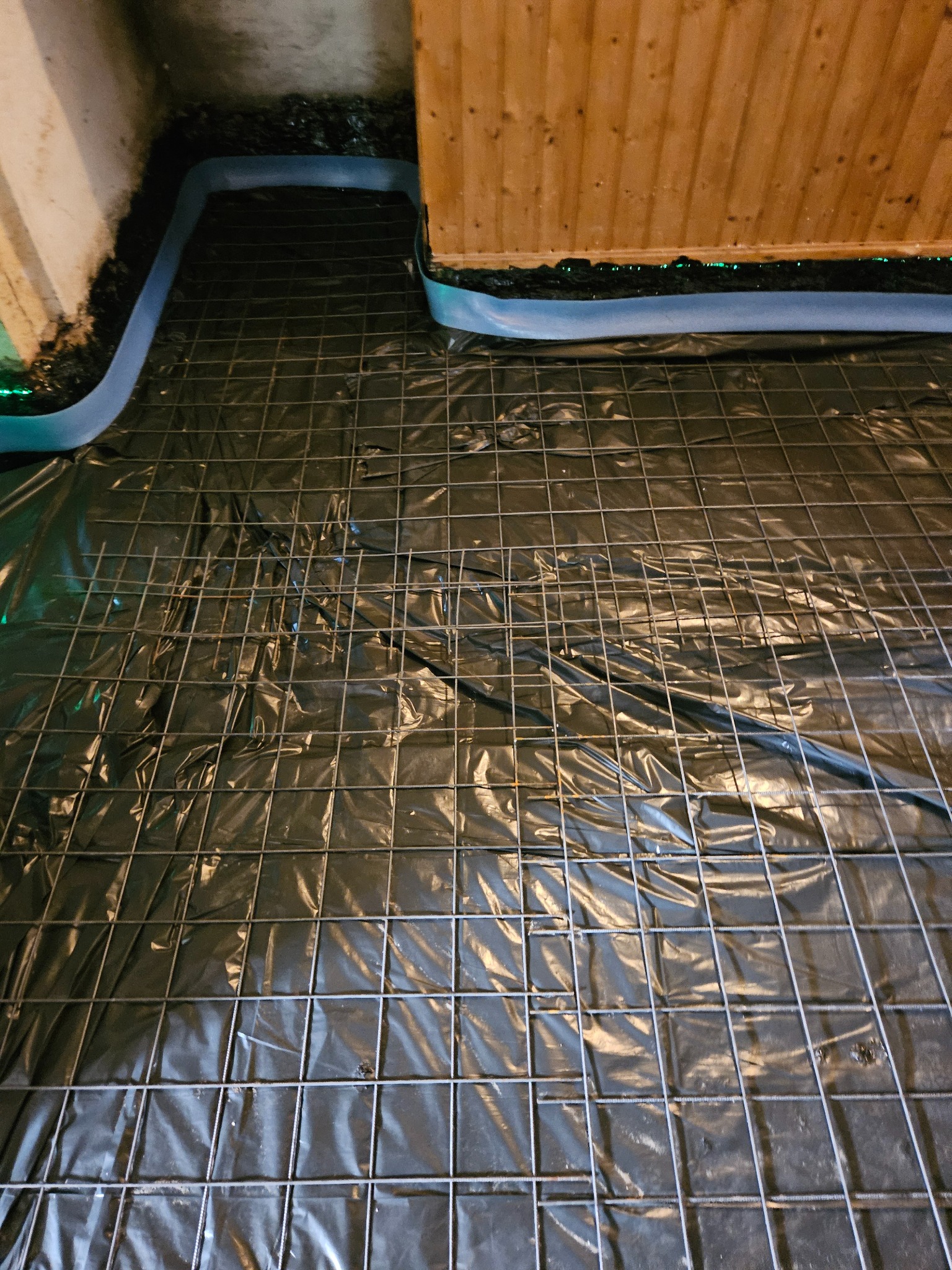 Przygotowanie podłoża: folia izolacyjna i siatka zbrojeniowa na podłożu, obrzeża zabezpieczone taśmą, widoczne fragmenty ściany i drewnianej konstrukcji.