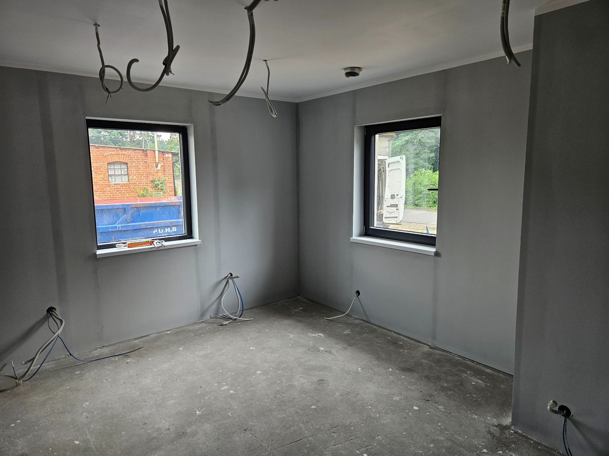 Szare, odświeżone wnętrze pokoju z dwoma oknami, widoczne wystające kable elektryczne ze ścian i sufitu przed montażem osprzętu.