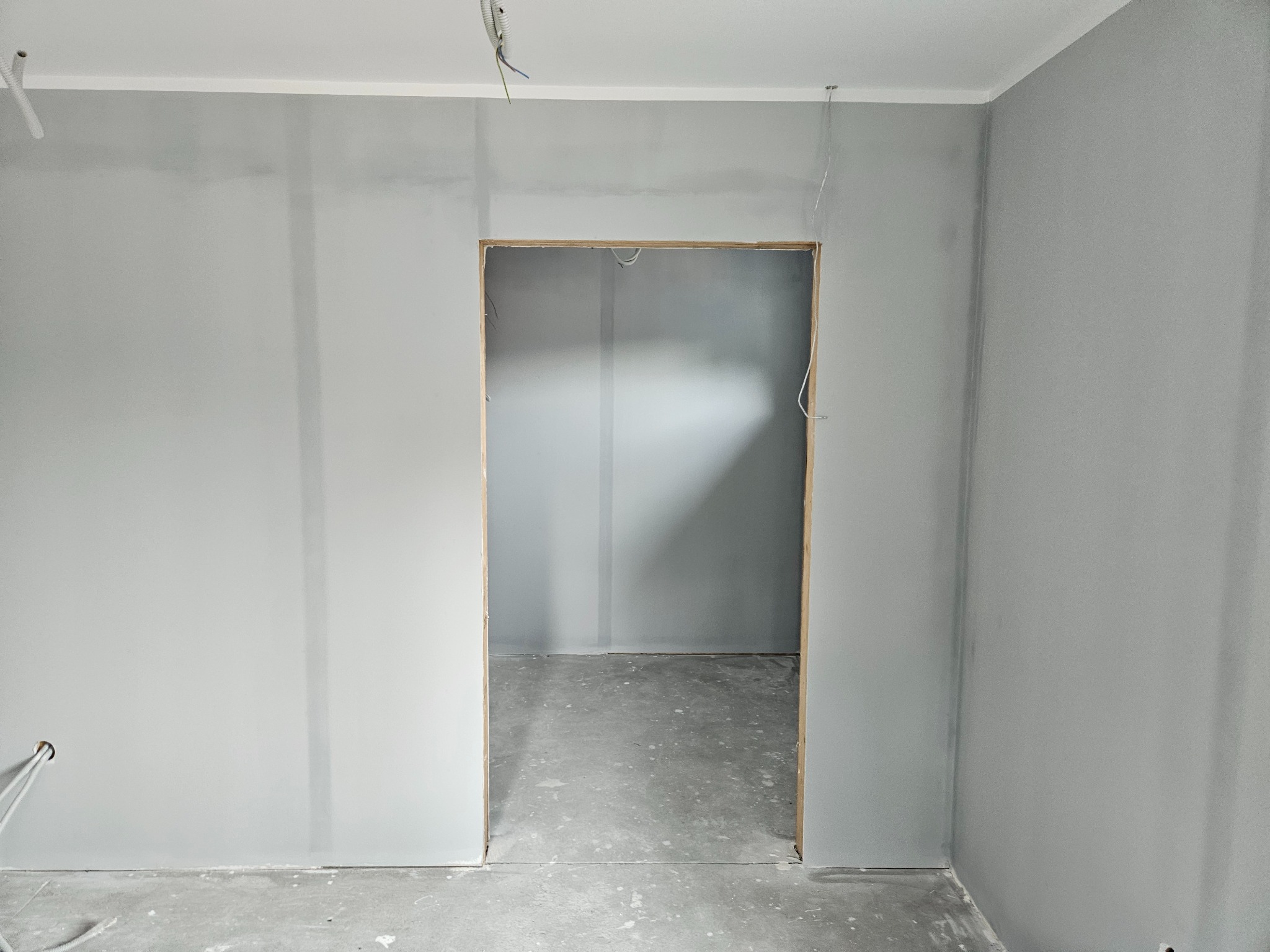 Przygotowane do malowania, zagruntowane ściany w szarym odcieniu, widoczne wystające kable elektryczne oraz surowy otwór drzwiowy bez ościeżnicy.