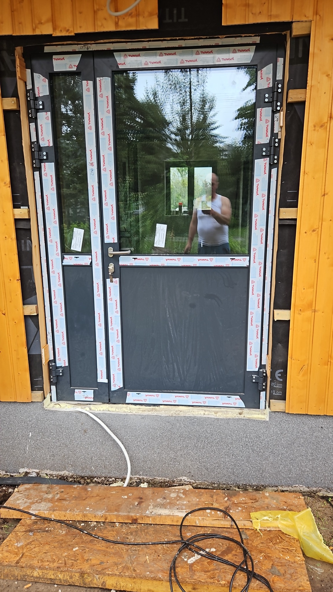 Montaż dwuskrzydłowych drzwi zewnętrznych z czarną ramą, widoczne tymczasowe zabezpieczenia i odblask sylwetki w szybie.