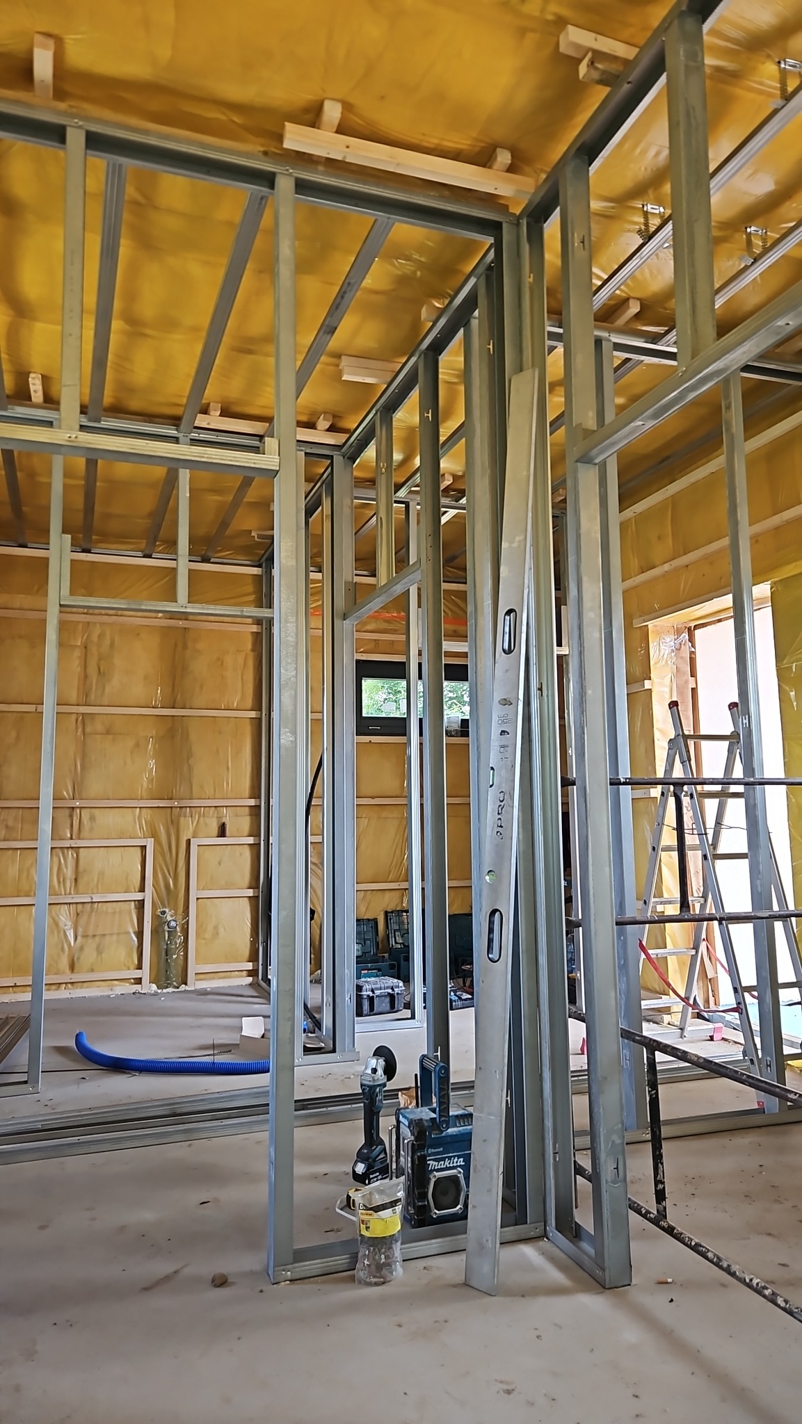 Wnętrze w trakcie prac wykończeniowych: widoczne metalowe profile ścian działowych, izolacja na ścianach i suficie, poziomica oparta o konstrukcję, narzędzia budowlane na podłodze.