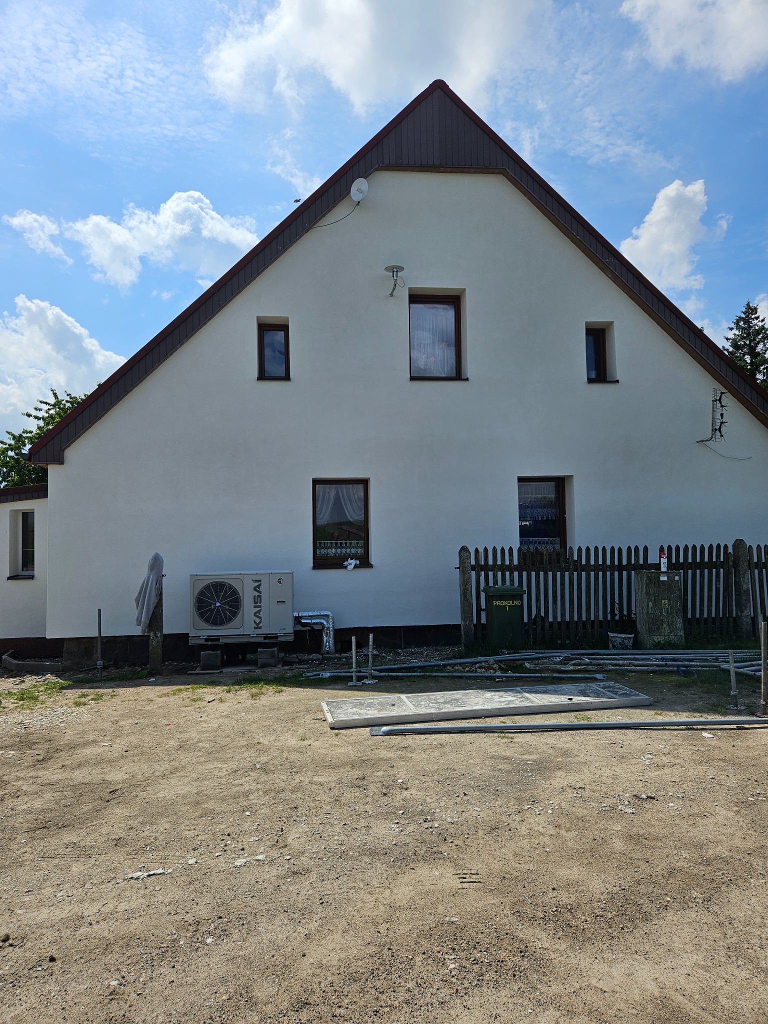 Biały dom jednorodzinny z brązowym dachem, widoczna jednostka zewnętrzna pompy ciepła KAISAI, drewniany płot i przygotowany teren pod prace ziemne.