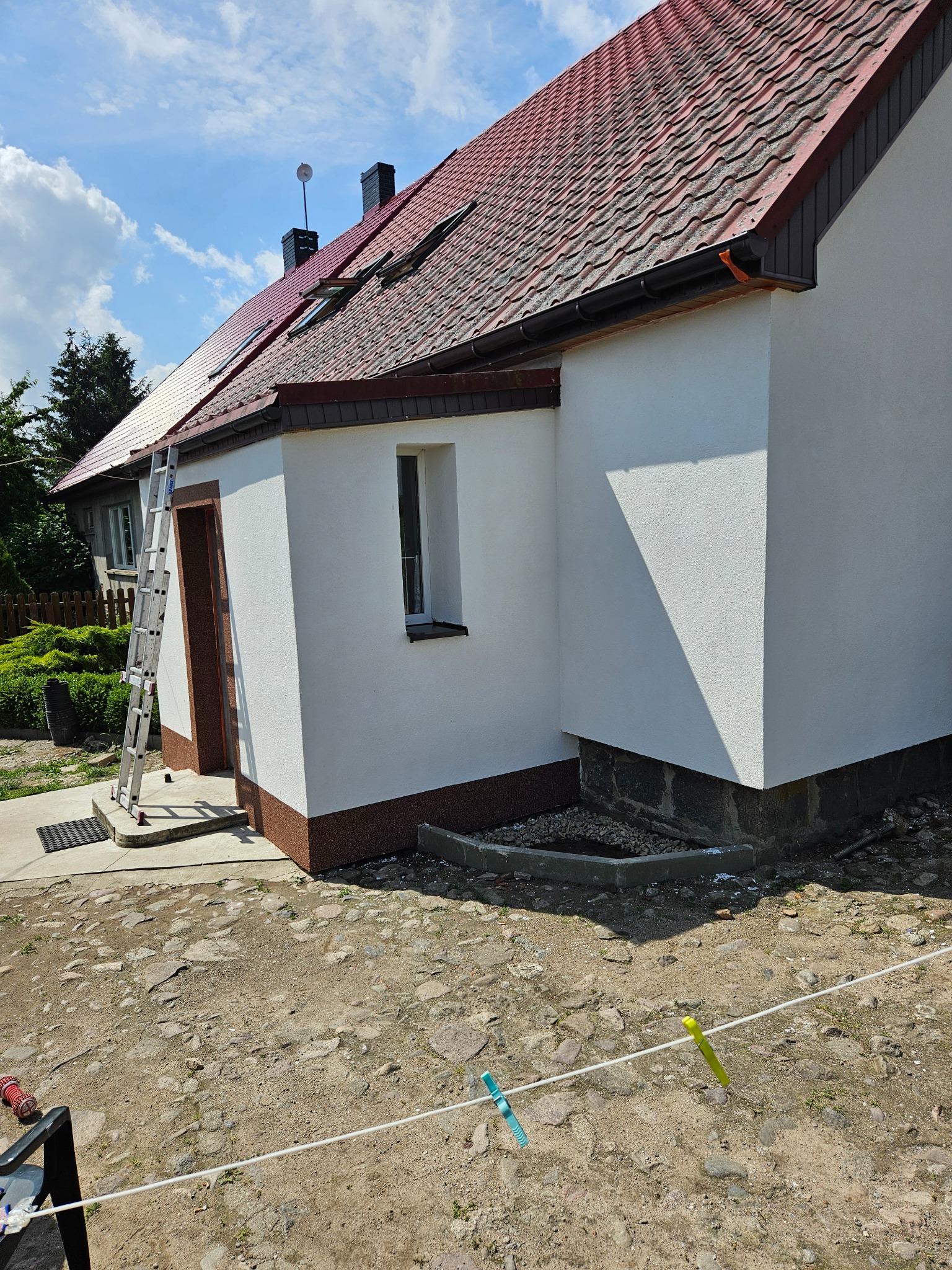 Odnowiona elewacja domu jednorodzinnego z nowym tynkiem i pomalowanymi elementami dekoracyjnymi, widoczna drabina oparta o ścianę, suszące się pranie na sznurku w pierwszym planie.