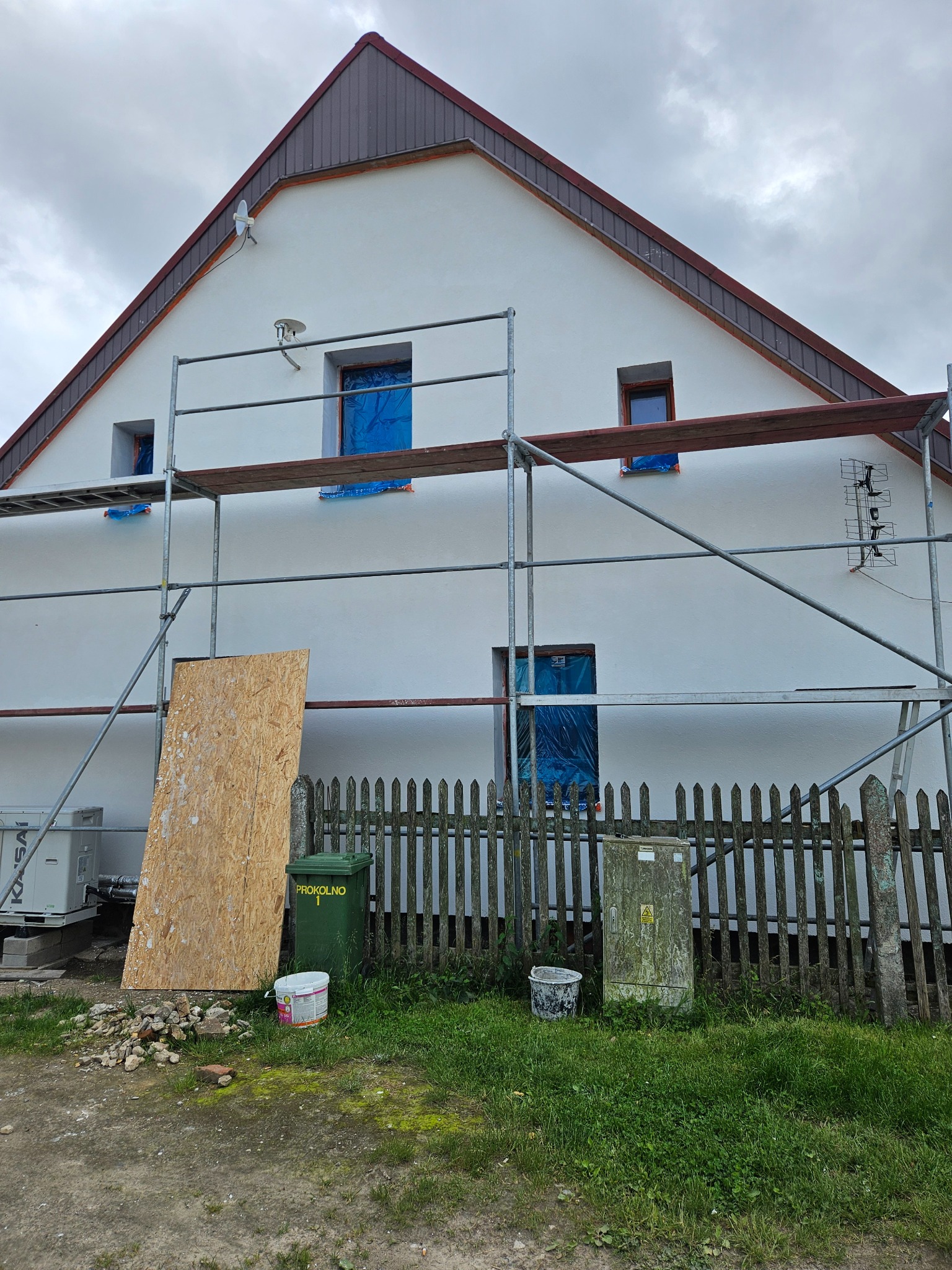 Dom jednorodzinny w trakcie prac elewacyjnych: rusztowanie, okna zabezpieczone folią, widoczne ślady po nałożonym tynku, niezabezpieczona antena na ścianie, przy płocie stoi tablica budowlana...