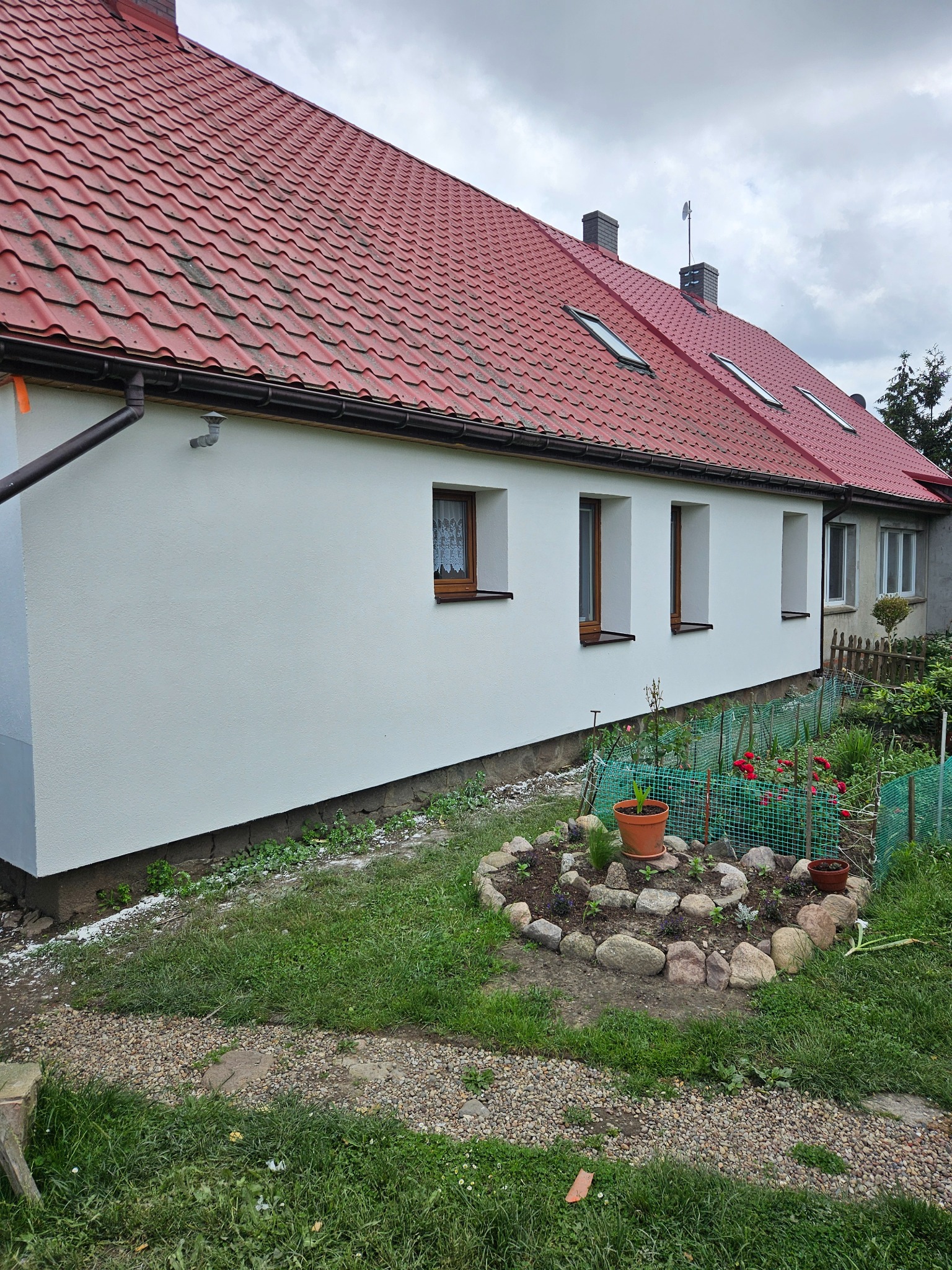 Odnowiona elewacja domu jednorodzinnego z czerwonym dachem, widok z boku, małe okna z drewnianymi ramami, ogródek z kwiatami i kamieniami wzdłuż ściany.