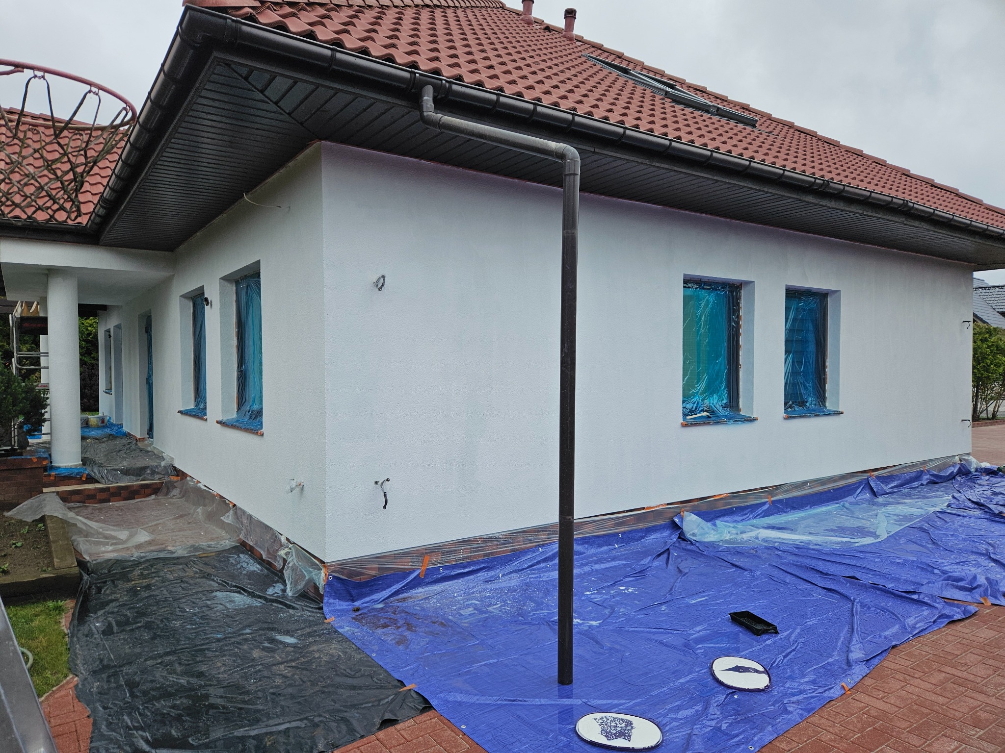 Biała elewacja domu jednorodzinnego w trakcie ocieplania, okna zabezpieczone folią, widoczne plandeki ochronne na ziemi i fragment dachu z czerwonej dachówki.