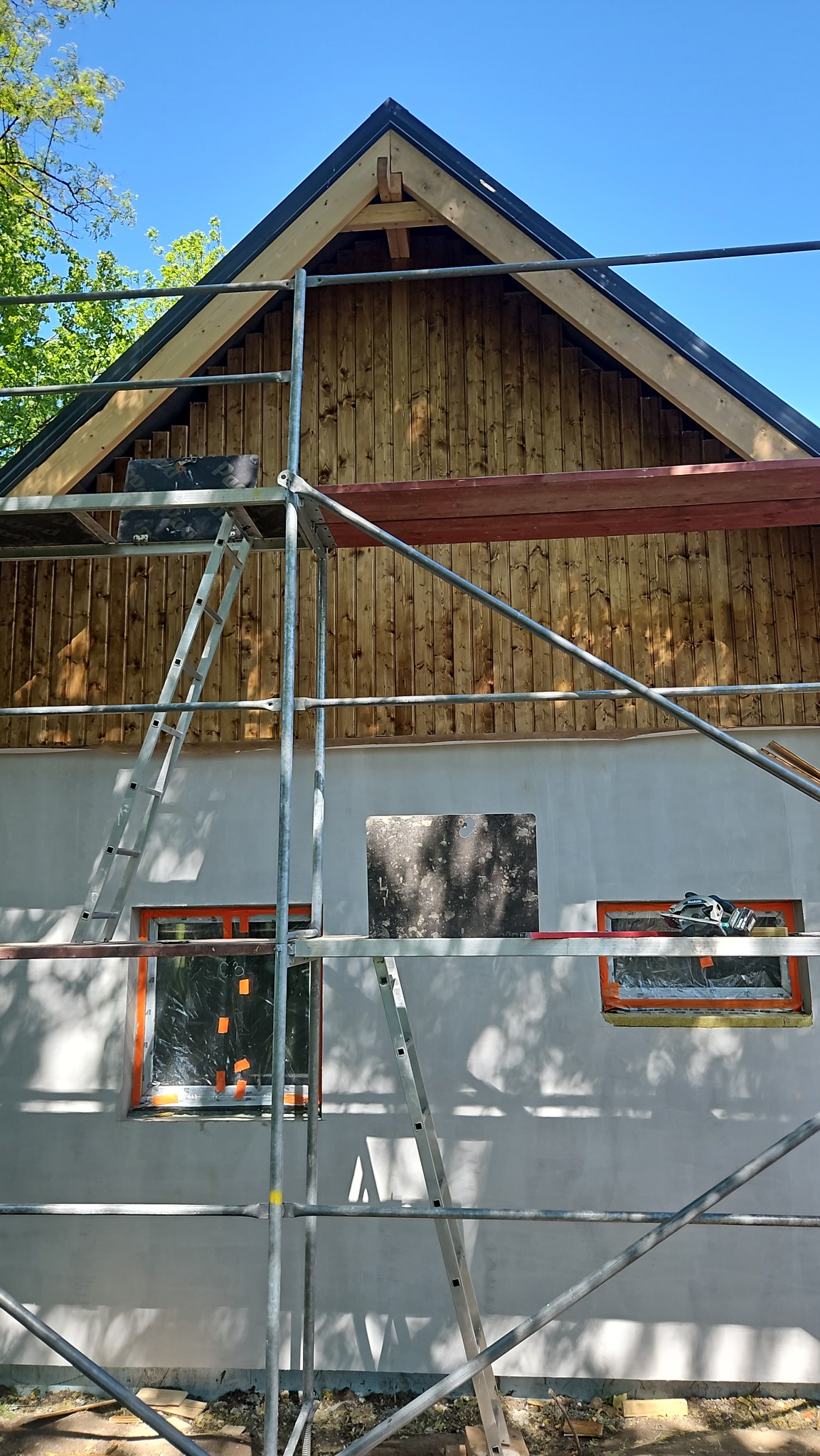 Dom w trakcie ocieplania elewacji, widoczne rusztowanie, ocieplona górna część ściany wykończona drewnem, okna zabezpieczone folią, przygotowane do dalszych prac.