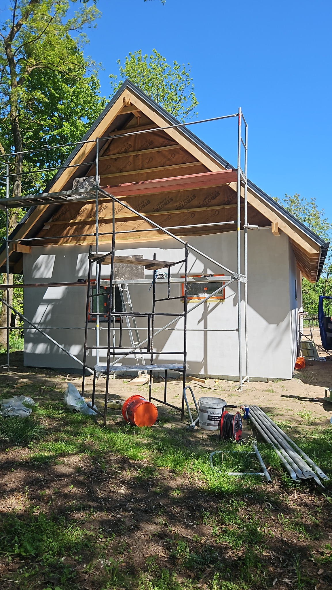 Budynek jednorodzinny w trakcie ocieplania elewacji, widoczne rusztowania, częściowo otynkowana ściana, odkryta konstrukcja dachu z membraną, narzędzia i materiały budowlane rozłożone na trawie.