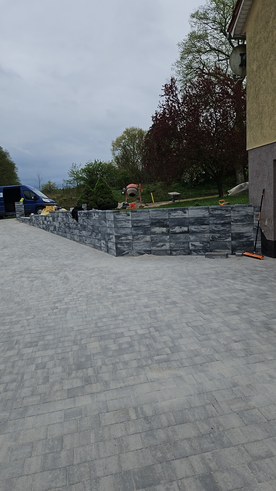 Szara kostka brukowa ułożona na podjeździe, z niskim murem oporowym z ciemniejszych kostek, w tle zaparkowany niebieski bus i betoniarka.
