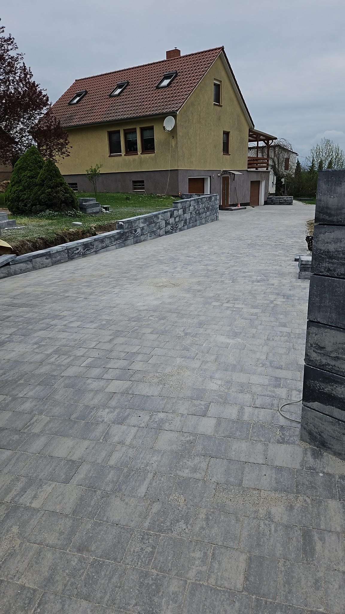 Ułożona szara kostka brukowa prowadząca do domu z beżową elewacją i czerwoną dachówką, z widocznym fragmentem murku z kostki i stosem nieułożonych elementów po prawej stronie.