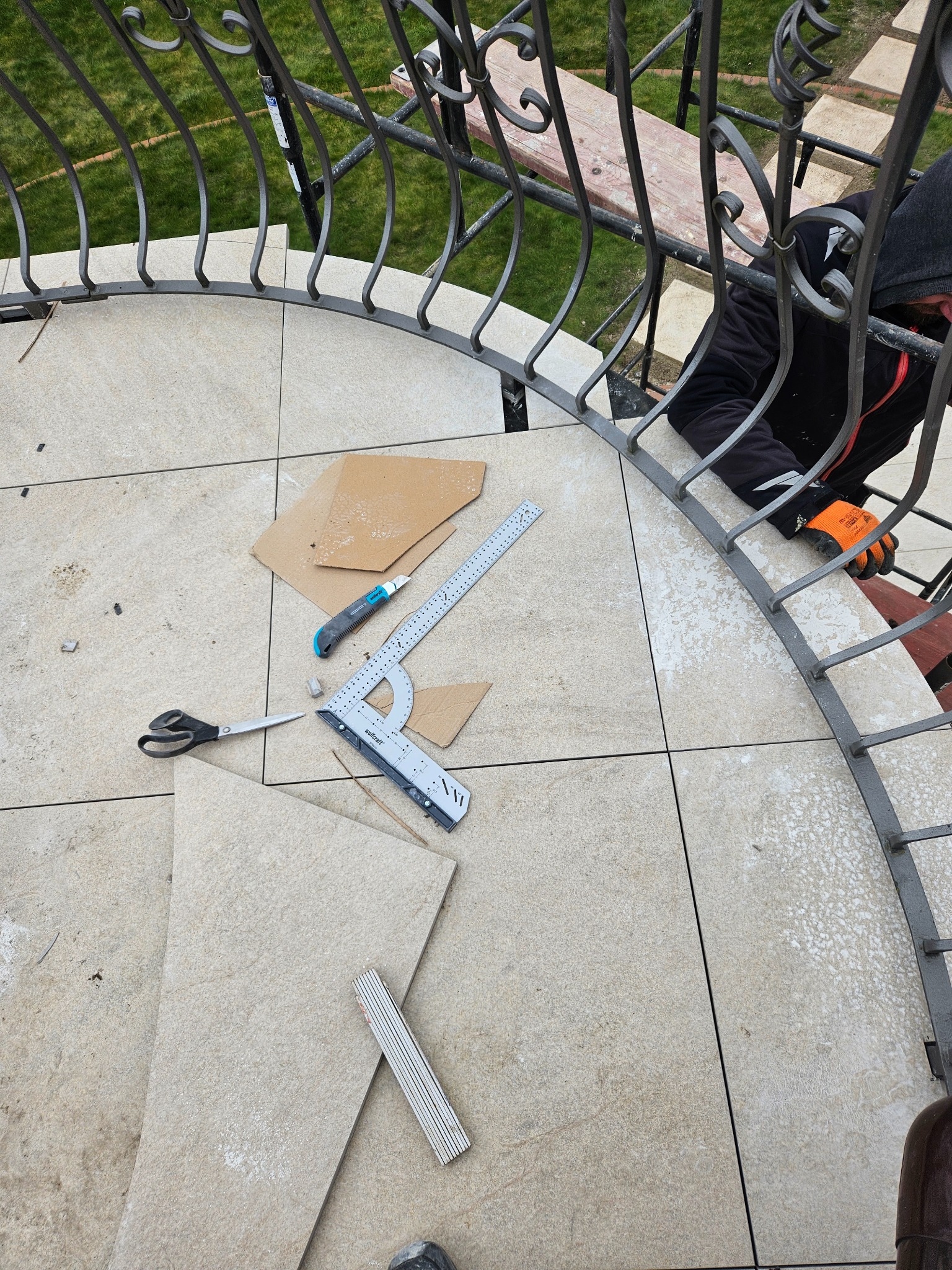 Montaż płytek na zakrzywionym balkonie z metalową balustradą; widoczne narzędzia: nóż, kątownik, nożyczki i miarka składana; pracownik w rękawicach ochronnych przycina płytkę.