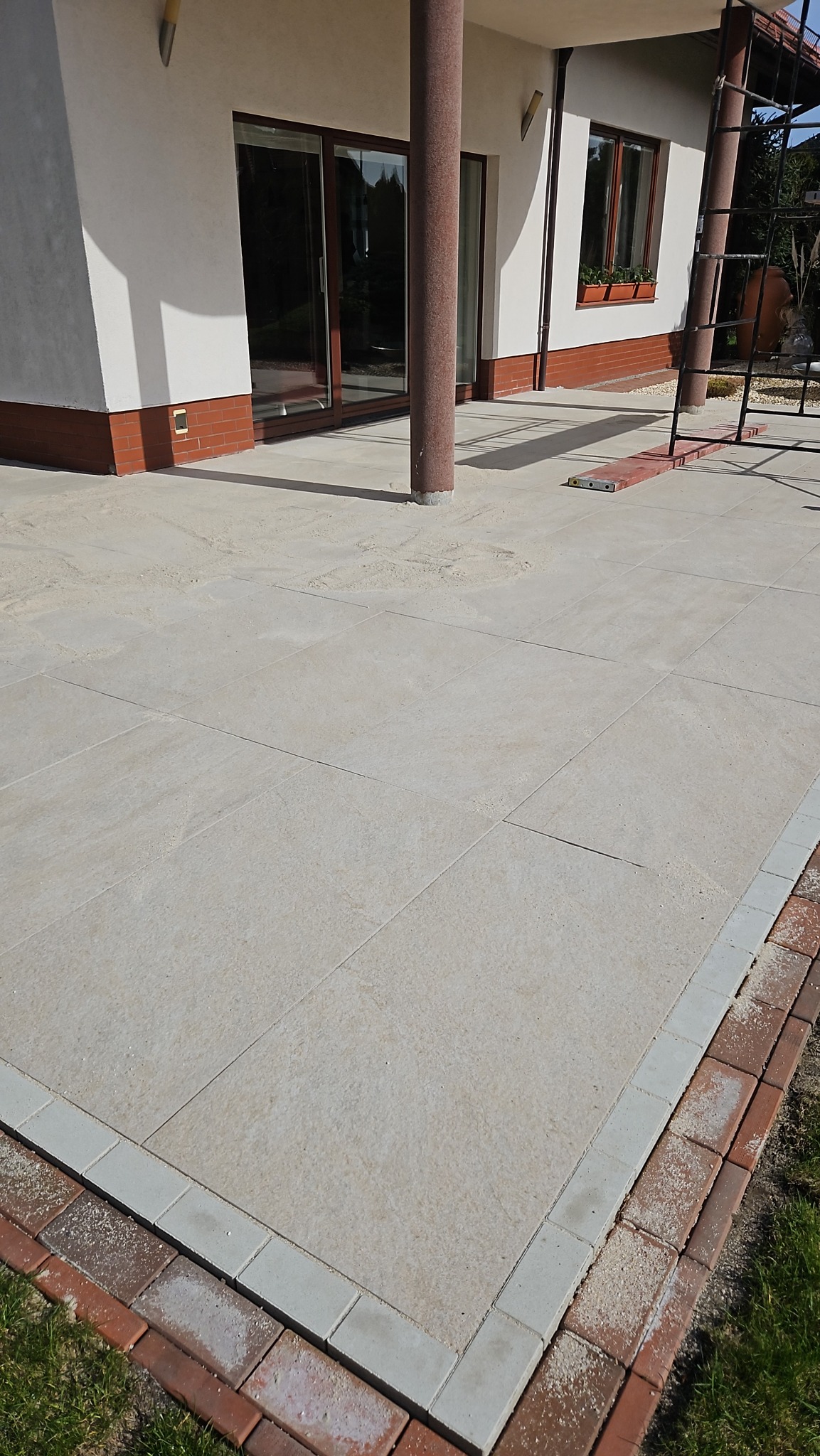Jasnoszara płyta tarasowa z obrzeżem z czerwonej cegły i szarej kostki, widok z góry na tle domu z brązowymi oknami i kolumną.