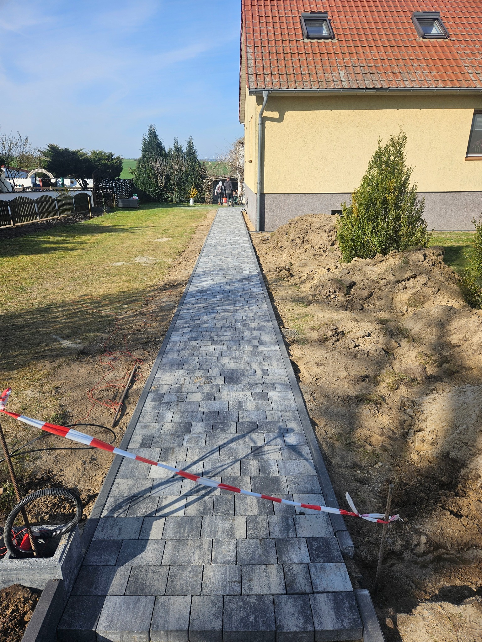 Świeżo ułożony chodnik z szarej kostki brukowej prowadzący wzdłuż domu z pomarańczowym dachem, zabezpieczony taśmą ostrzegawczą, z widoczną stertą ziemi i dwoma osobami w oddali.