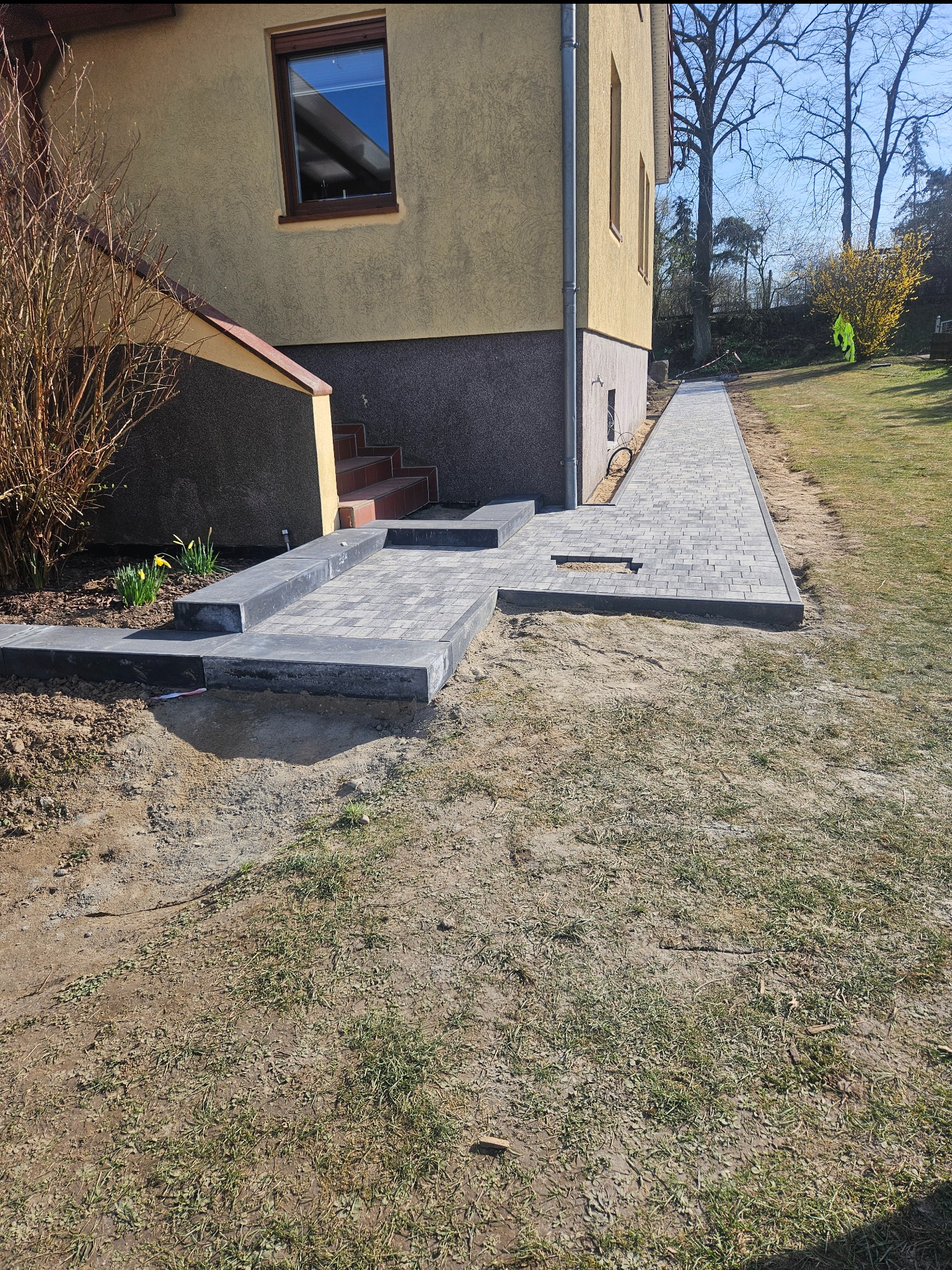 Świeżo ułożona, szara kostka brukowa tworząca chodnik i mały placyk przed wejściem do domu z żółtą elewacją. Widoczne niedociągnięcia w postaci piasku na kostce i brakującej kostki w chodniku.