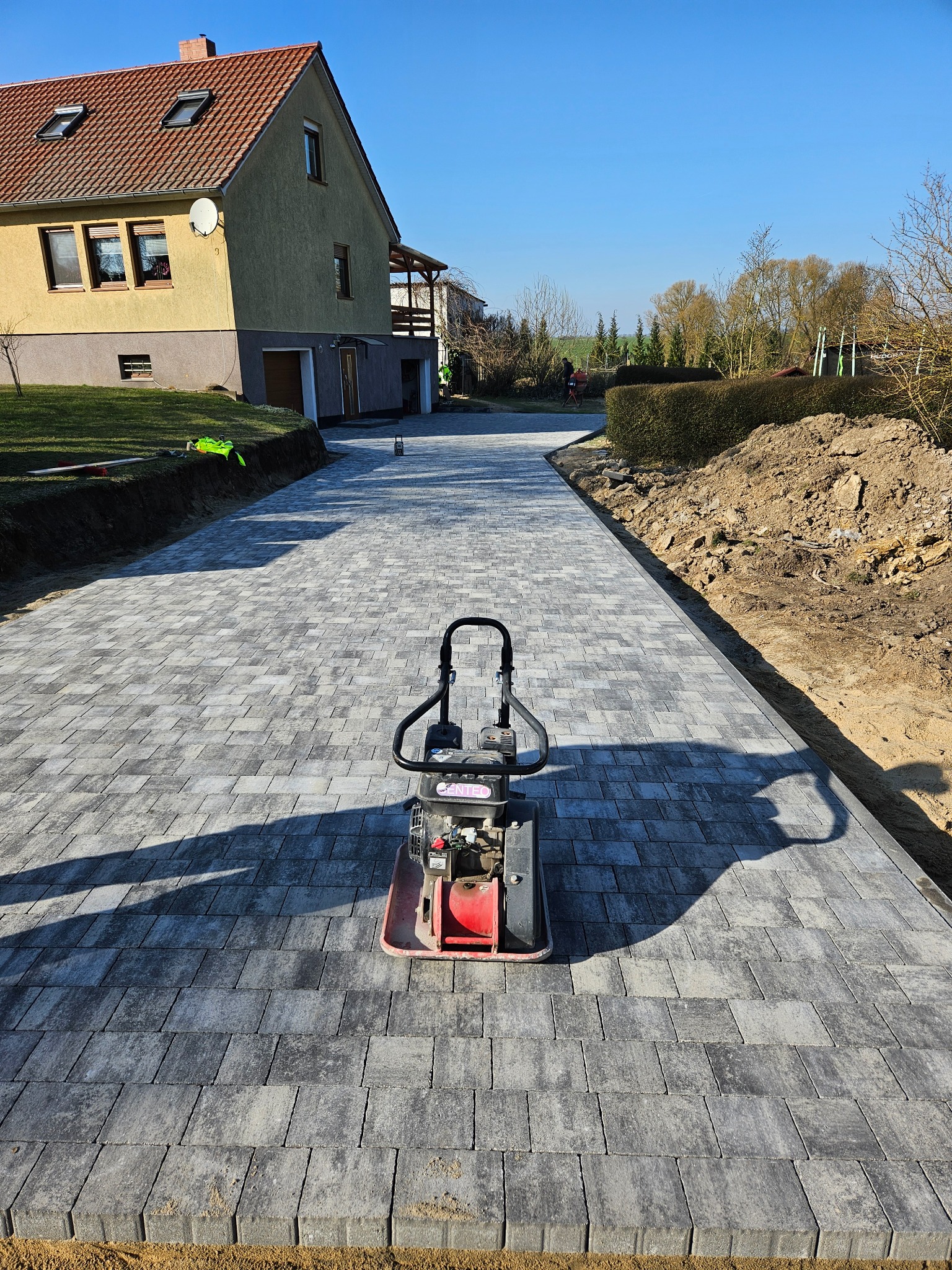 Świeżo ułożona, szara kostka brukowa prowadząca do domu, z zagęszczarką w centrum kadru i widoczną ziemią po bokach ścieżki.