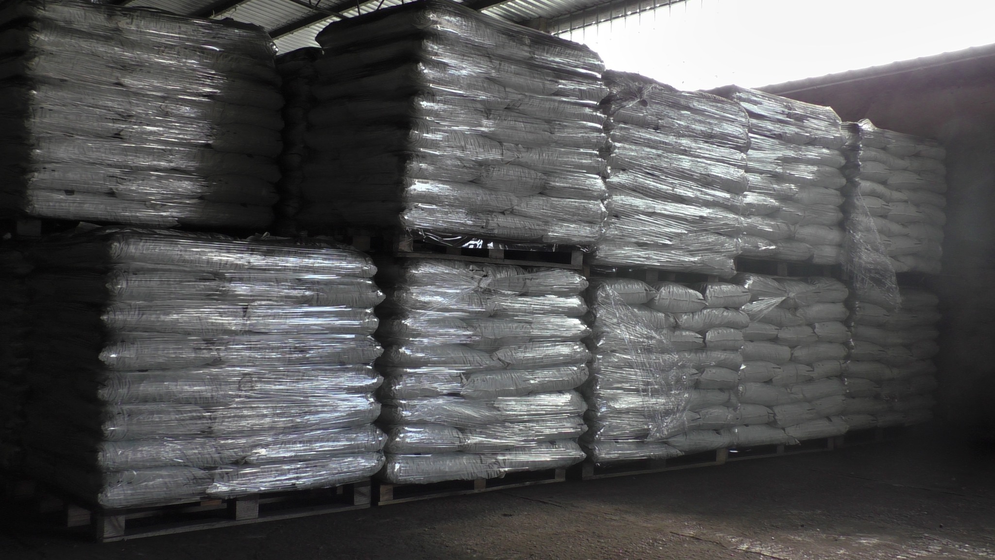 Stosy worków z opałem ułożone na paletach, zabezpieczone folią stretch w magazynie z metalową konstrukcją dachu i słabym oświetleniem.