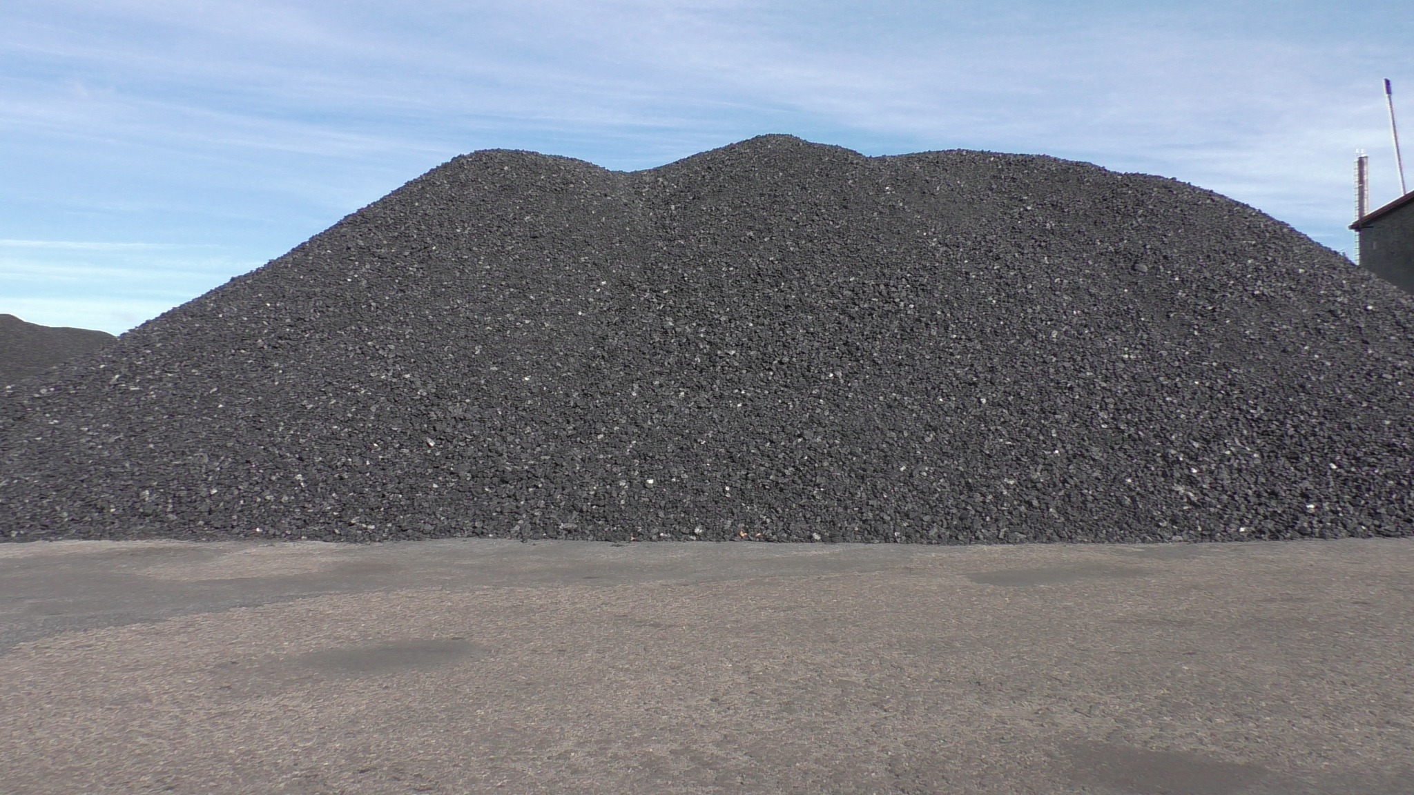 Góra czarnego węgla na betonowym placu, widoczne fragmenty budynku przemysłowego i niebo z delikatnymi chmurami.