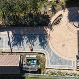 ECO FARM TECH SPÓŁKA Z OGRANICZONĄ ODPOWIEDZIALNOŚCIĄ - Widok z góry na posesję z nowo ułożoną kostką brukową w odcieniach szarości i beżu, tworzącą wzory geometryczne, widoczny traktor i elementy ogrodzenia w tle.