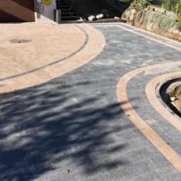 Podjazd z kostki brukowej w kolorach szarości i beżu, ułożony w łukowate wzory, z widoczną studzienką kanalizacyjną i obrzeżami, obok trawnik z roślinnością.