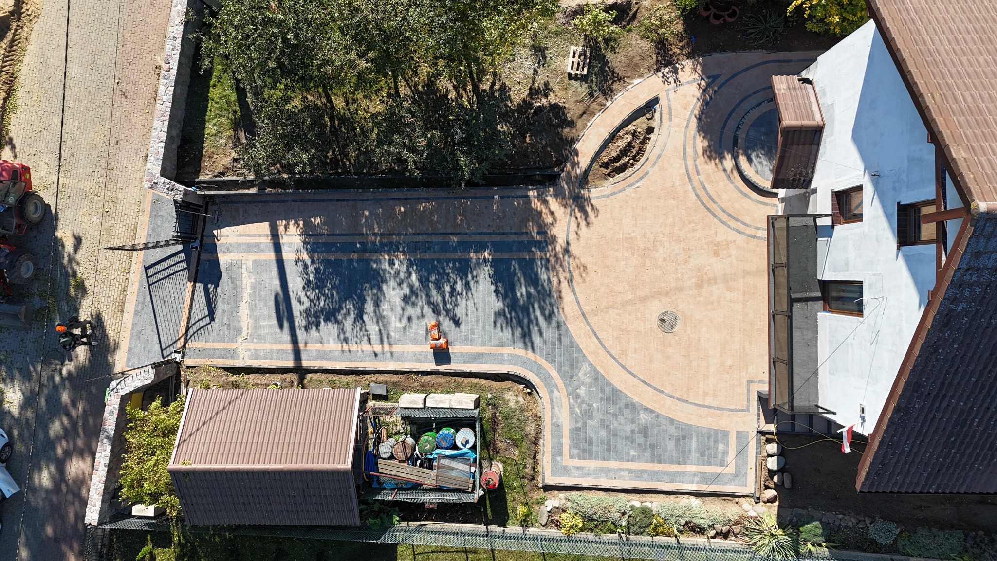 Widok z góry na posesję z nowo ułożoną kostką brukową w odcieniach szarości i beżu, tworzącą wzory geometryczne, widoczny traktor i elementy ogrodzenia w tle.