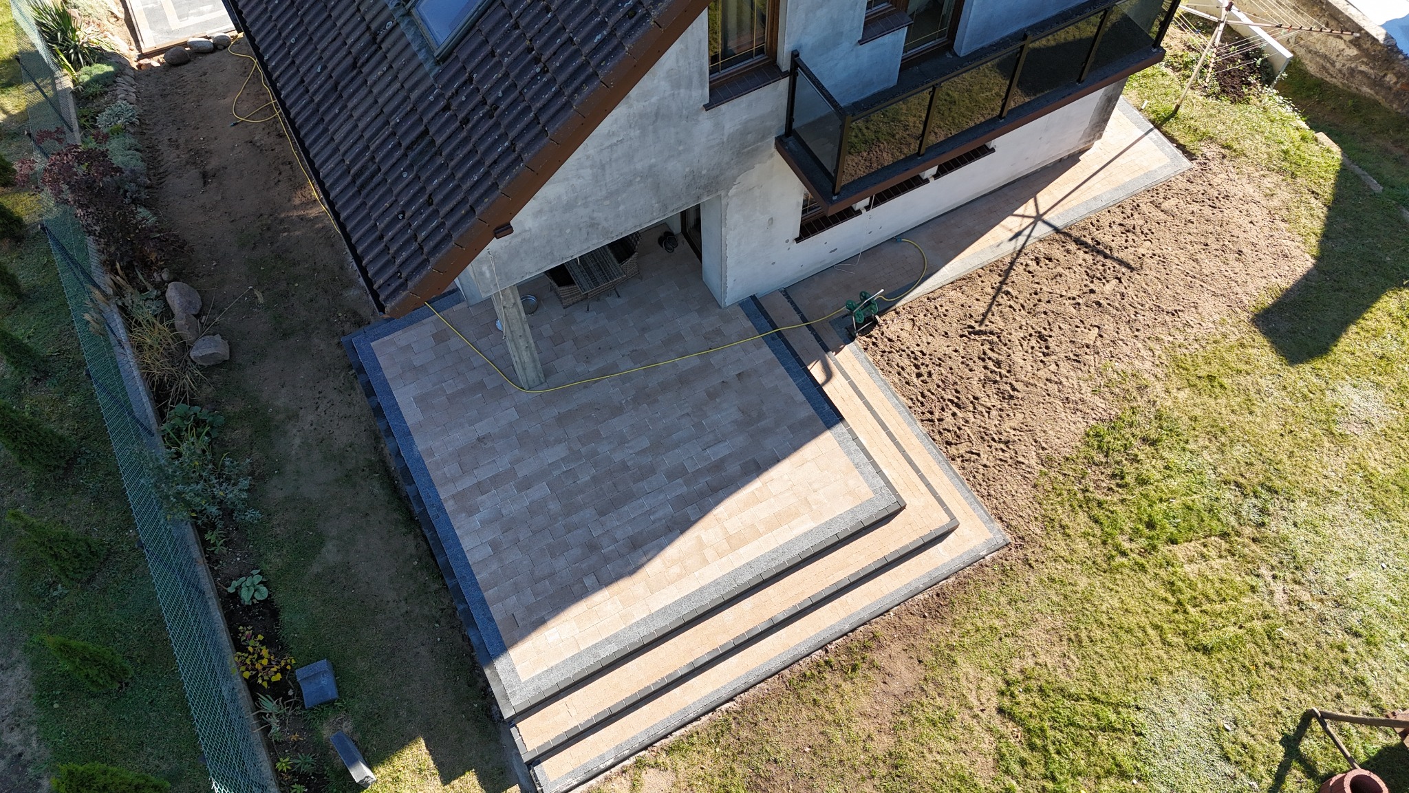 Widok z góry na nowo wyłożony taras z kostki brukowej z trzema stopniami, obok przygotowany teren pod trawnik i fragment domu z balkonem.